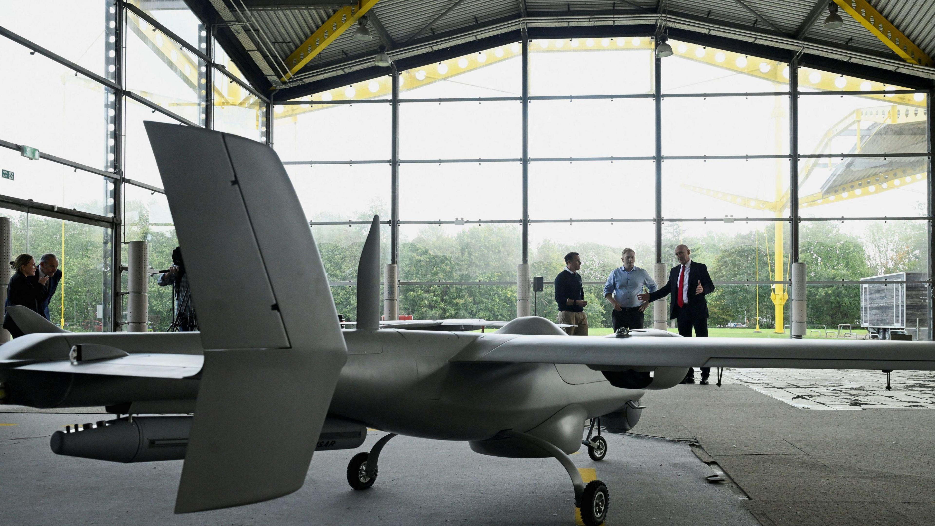 Shot taken from inside the building behind one of the drones, which looks like a miniature grey aeroplane. Three men speak behind it with the large glass panels of the building behind them