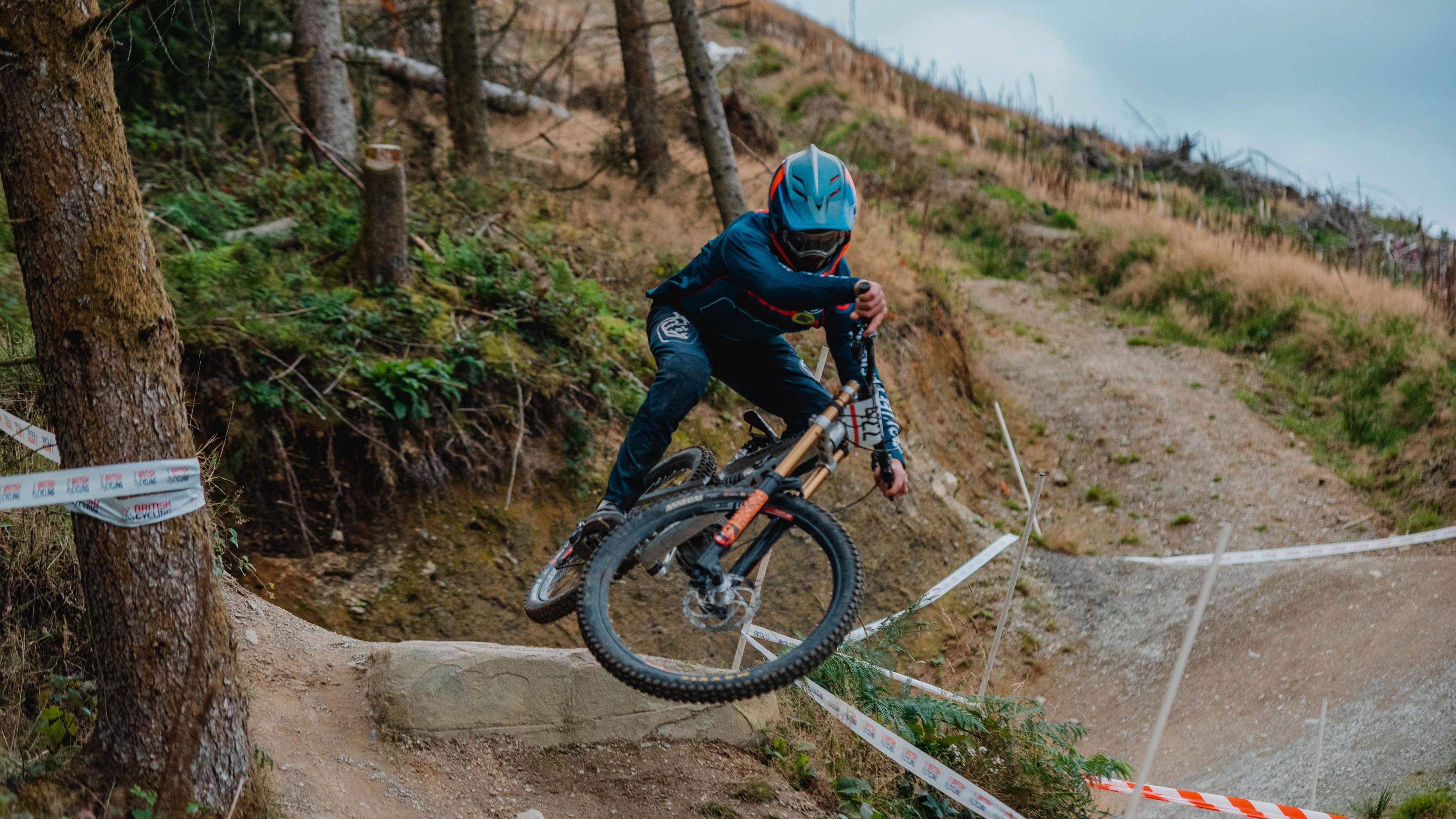 Ioan Hepburn in his teens competing in a downhill mountain bike race. The location is forestry with a twisty rutted track going between the trees, some of which have been felled.
He's taking a jump on his bike which is turned almost completely on its side in the air, and is wearing a light blue helmet.