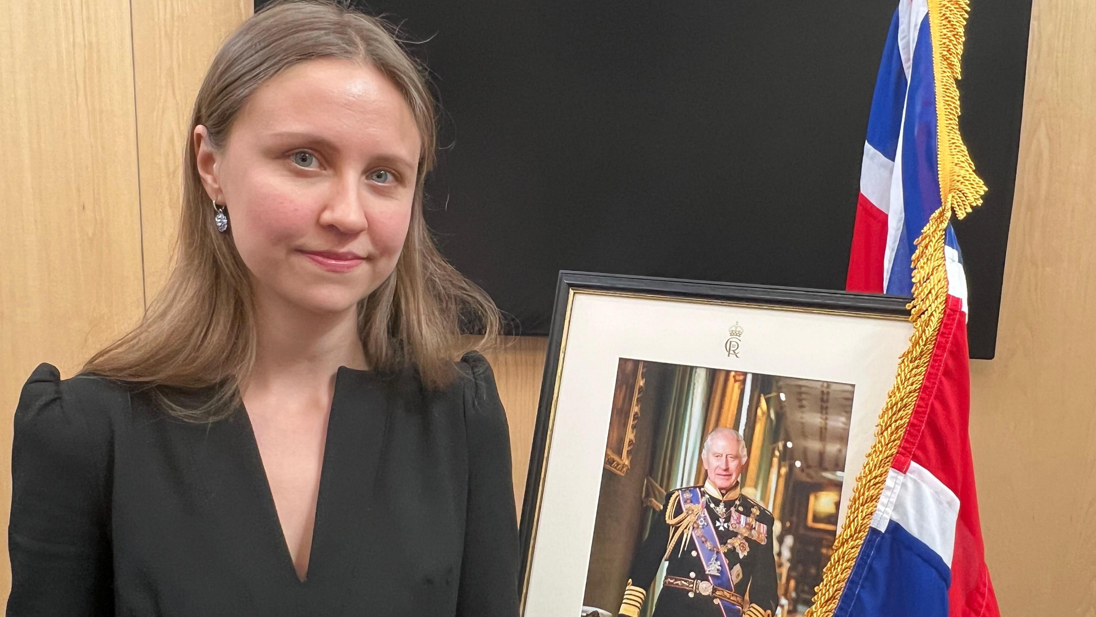 A young woman with shoulder-length fair hair, wearing a black dress, looks directly at the camera. She is next to a picture of the King, which is draped by a union jack.