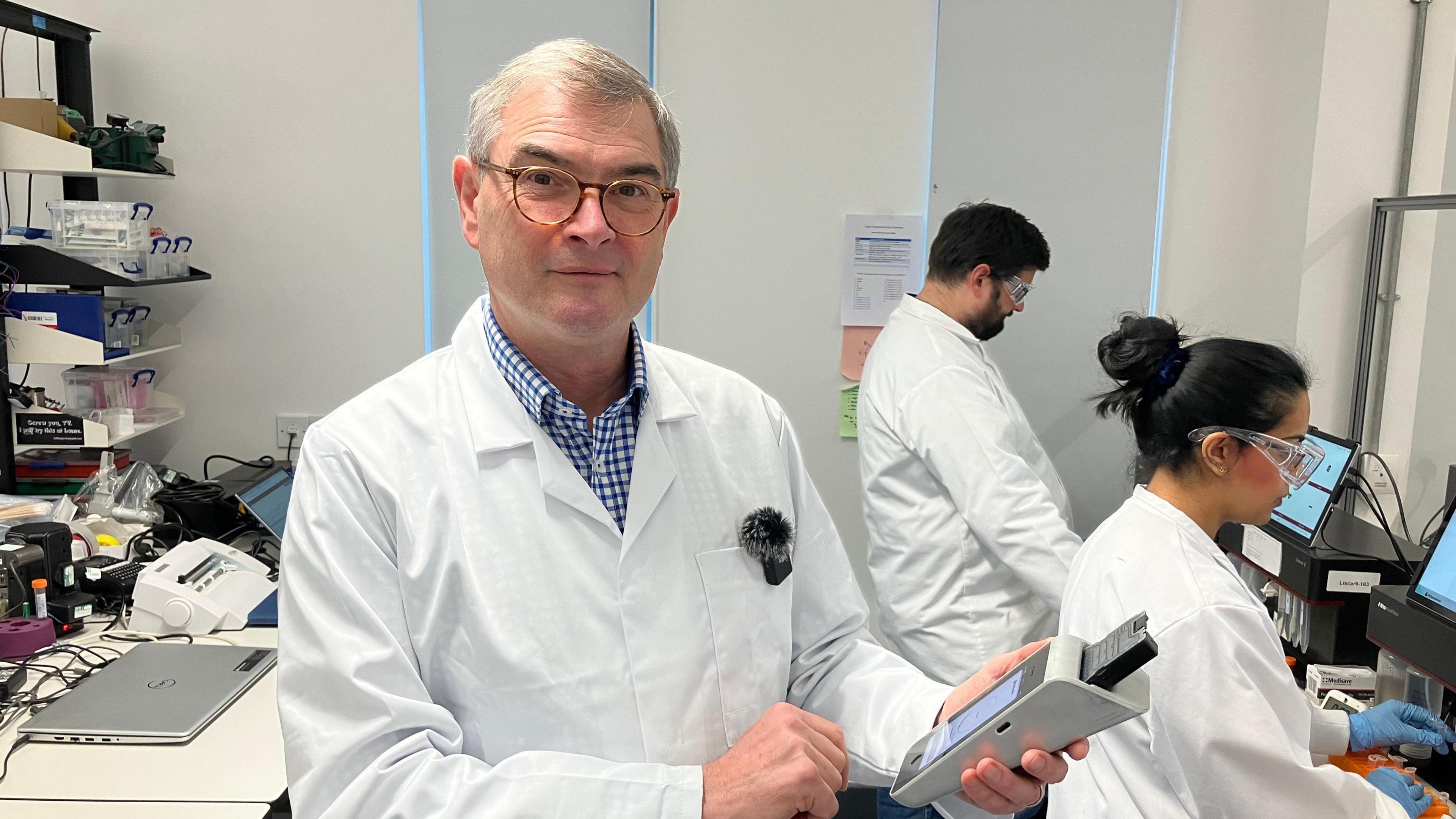 A man in a white laboratory coat, with greying hair and glasses , holding a testing machine in front of a set of researchers overlooking medical equipment.