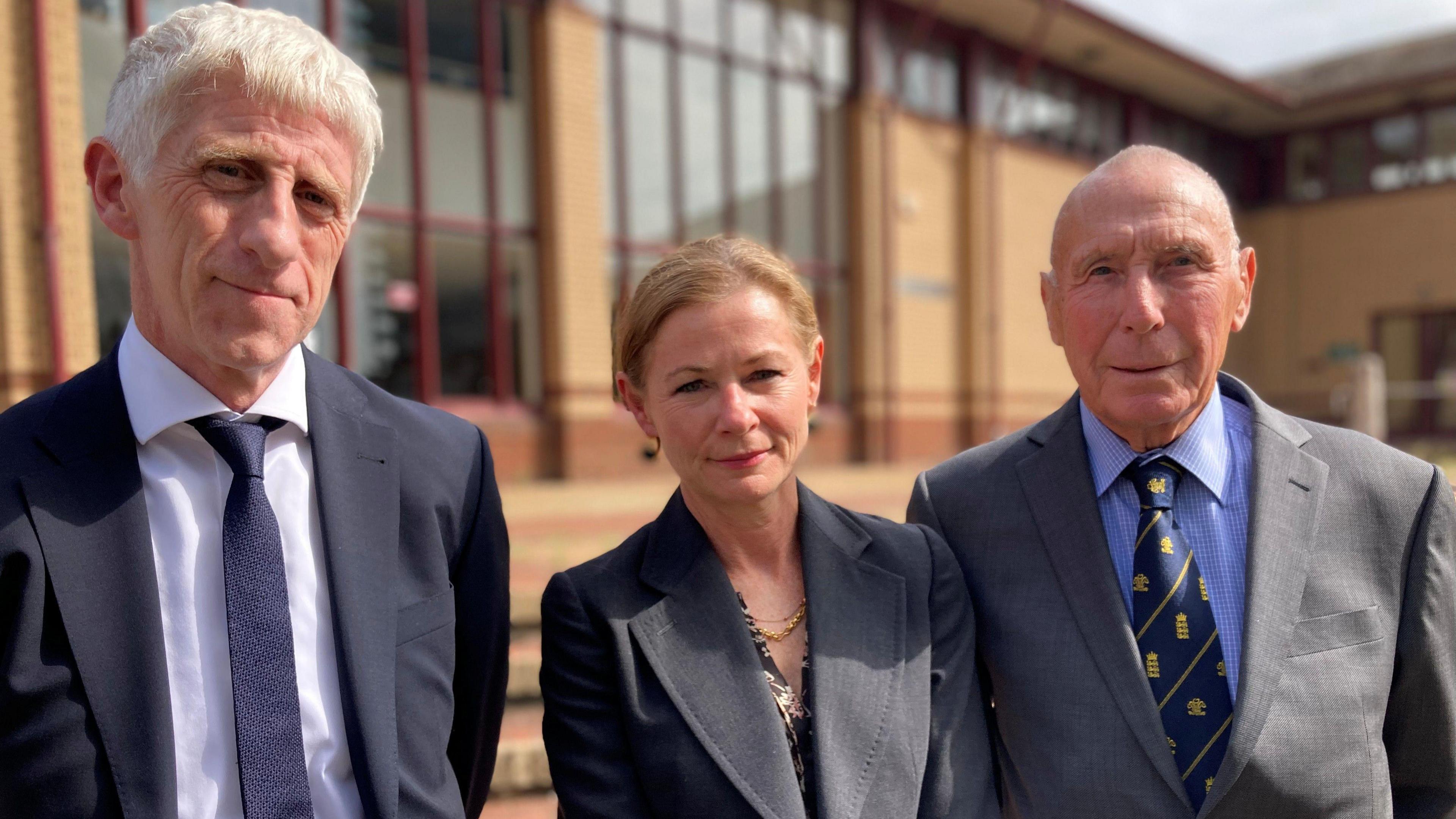 A man in a black suit and tie, standing next to a woman in a dark suit and she is standing next to another man wearing a dark grey suit. They are standing in front of Surrey Coroner's Court.