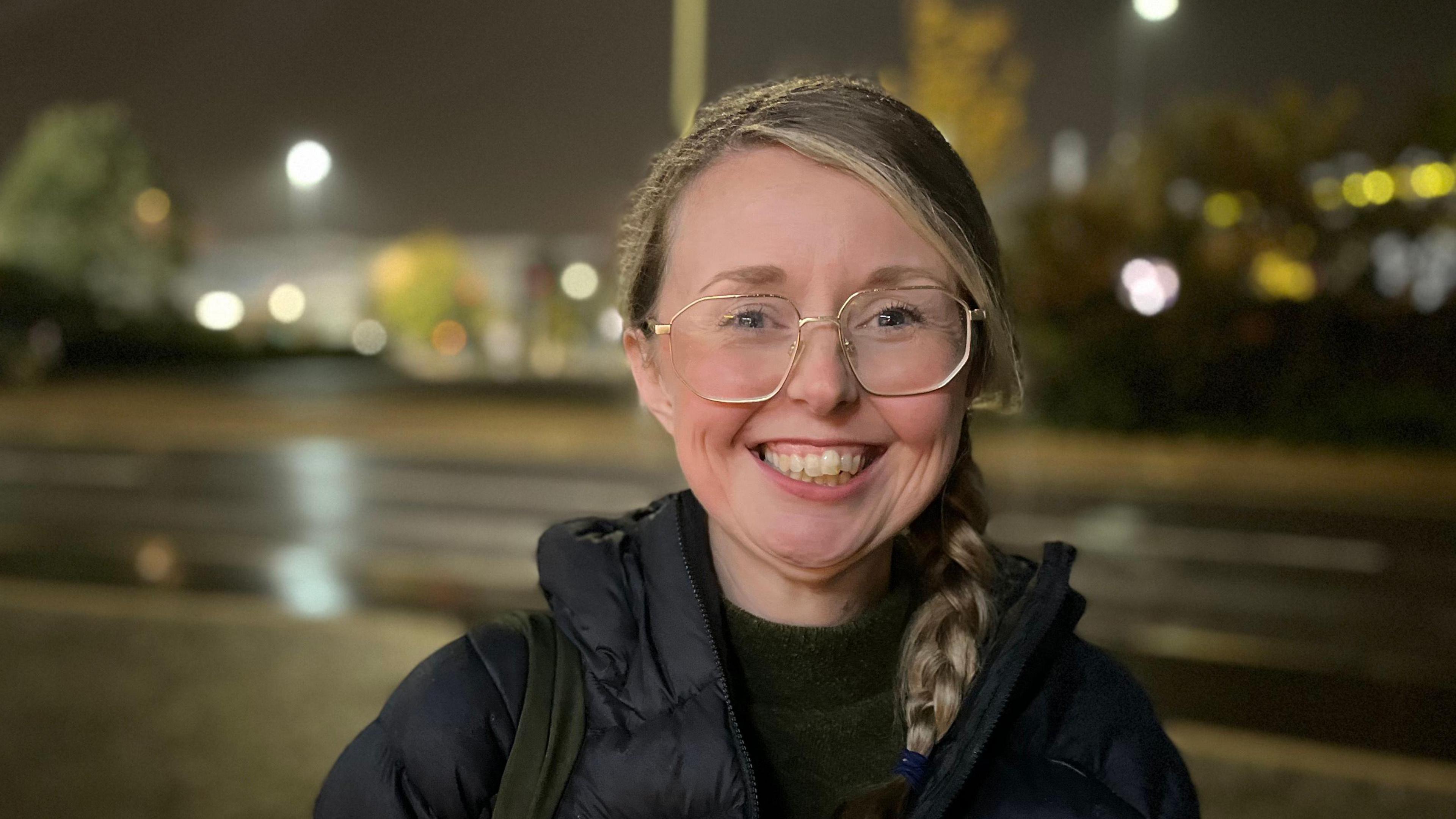 A woman in a black padded jacket. She is wearing glasses and has long blonde hair parted at the side and in a plait, which has been brought forward on to her left shoulder. She is standing outside at night beside a road which appears wet after rain.