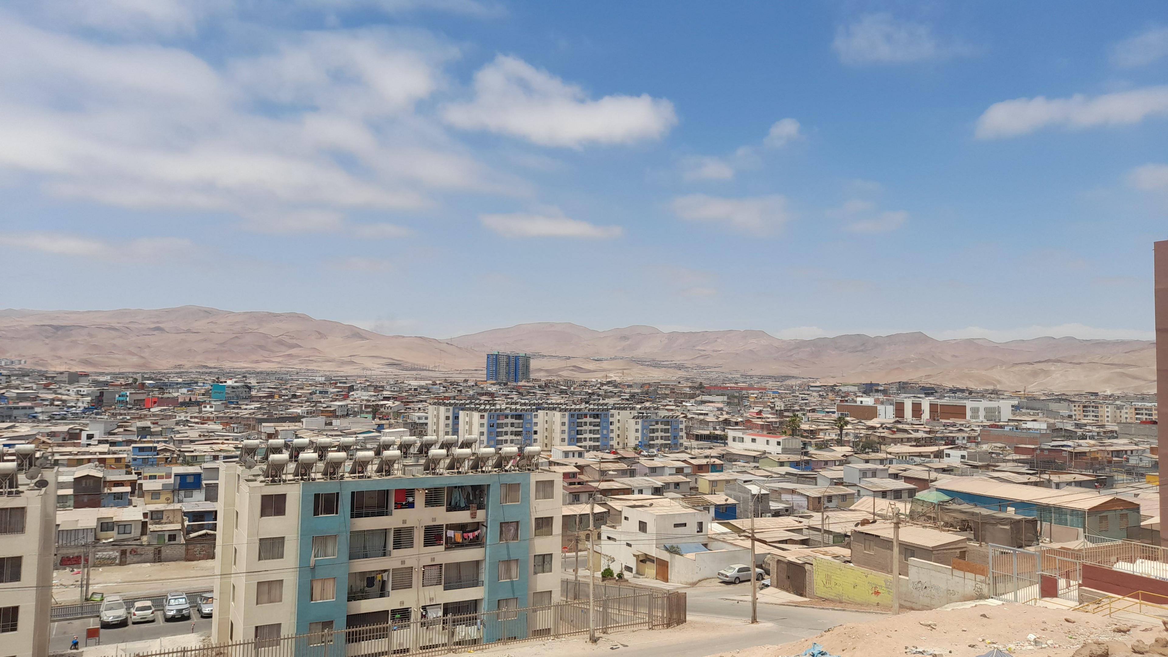 The image shows a slum, or informal settlement of shacks, in the Chilean city of Alto Hospico. There is a dense collection of low level buildings in an arid environment with desert mountains in the background