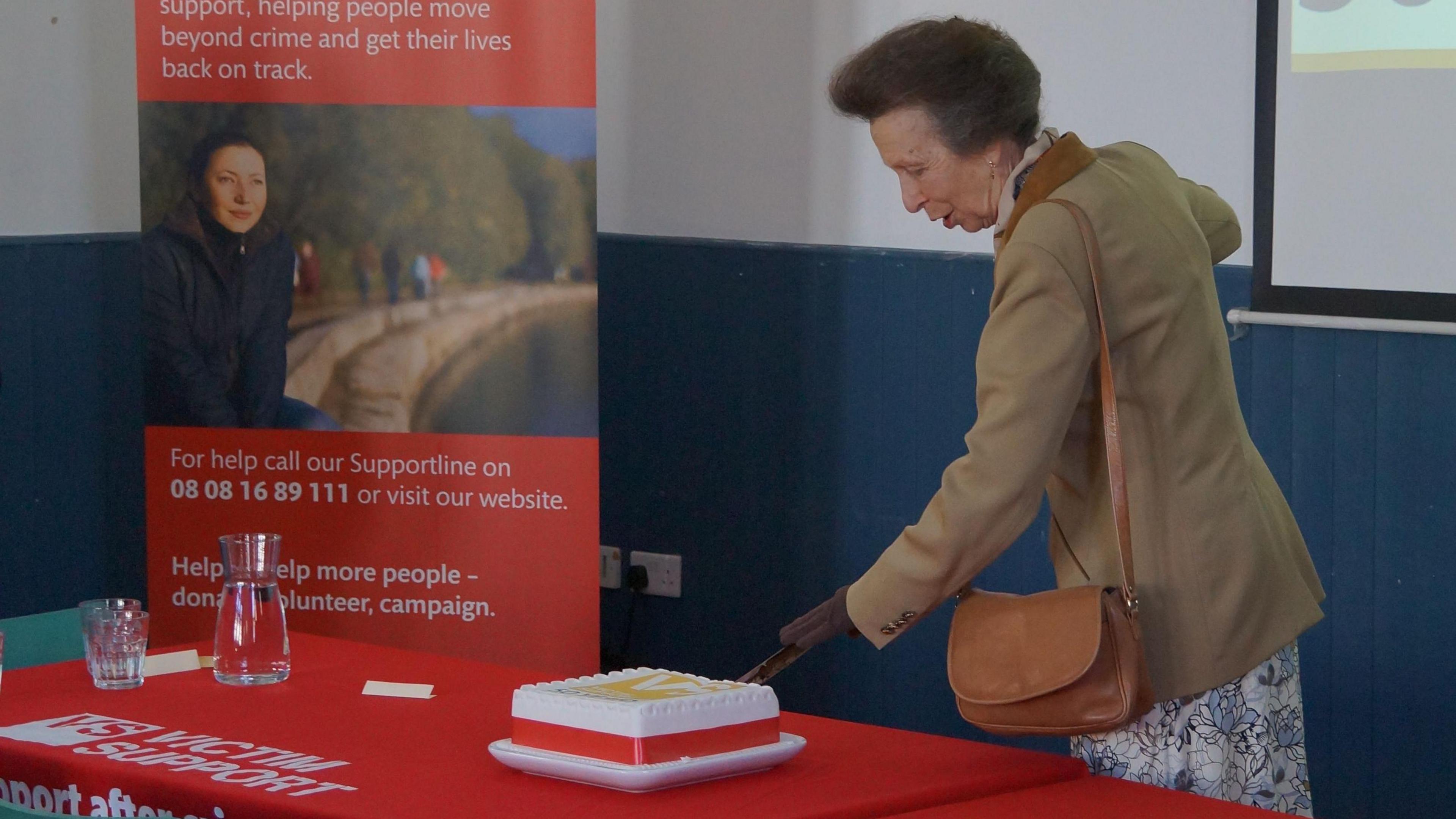 Princess Anne wearing a camel-coloured blazer, brown gloves and brown leather purse. She is cutting a white rectangular cake on a table in front of her using a long sword. In the background there is a Victim Support banner signposting where to go for help. 