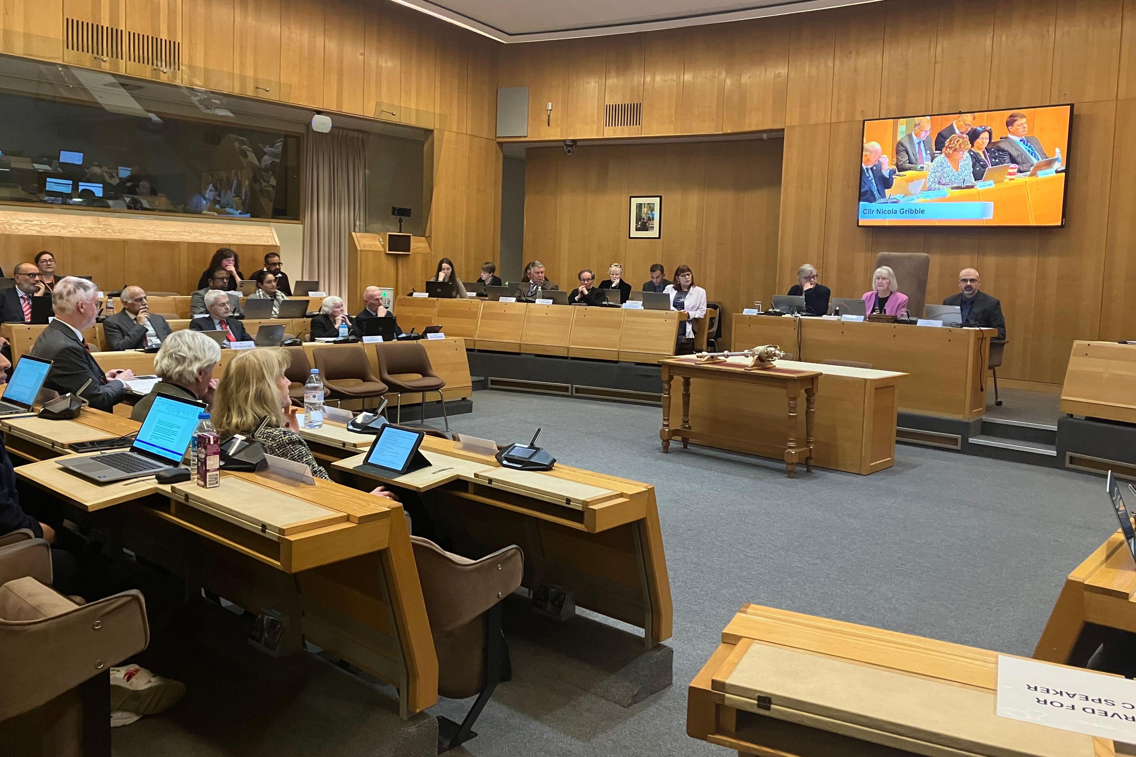 A picture of a room full of elected councillors who are in session and discussing the East West Rail project. It is a modern refurbished council chamber, with a big screen fixed to the wall about the chairperson's head