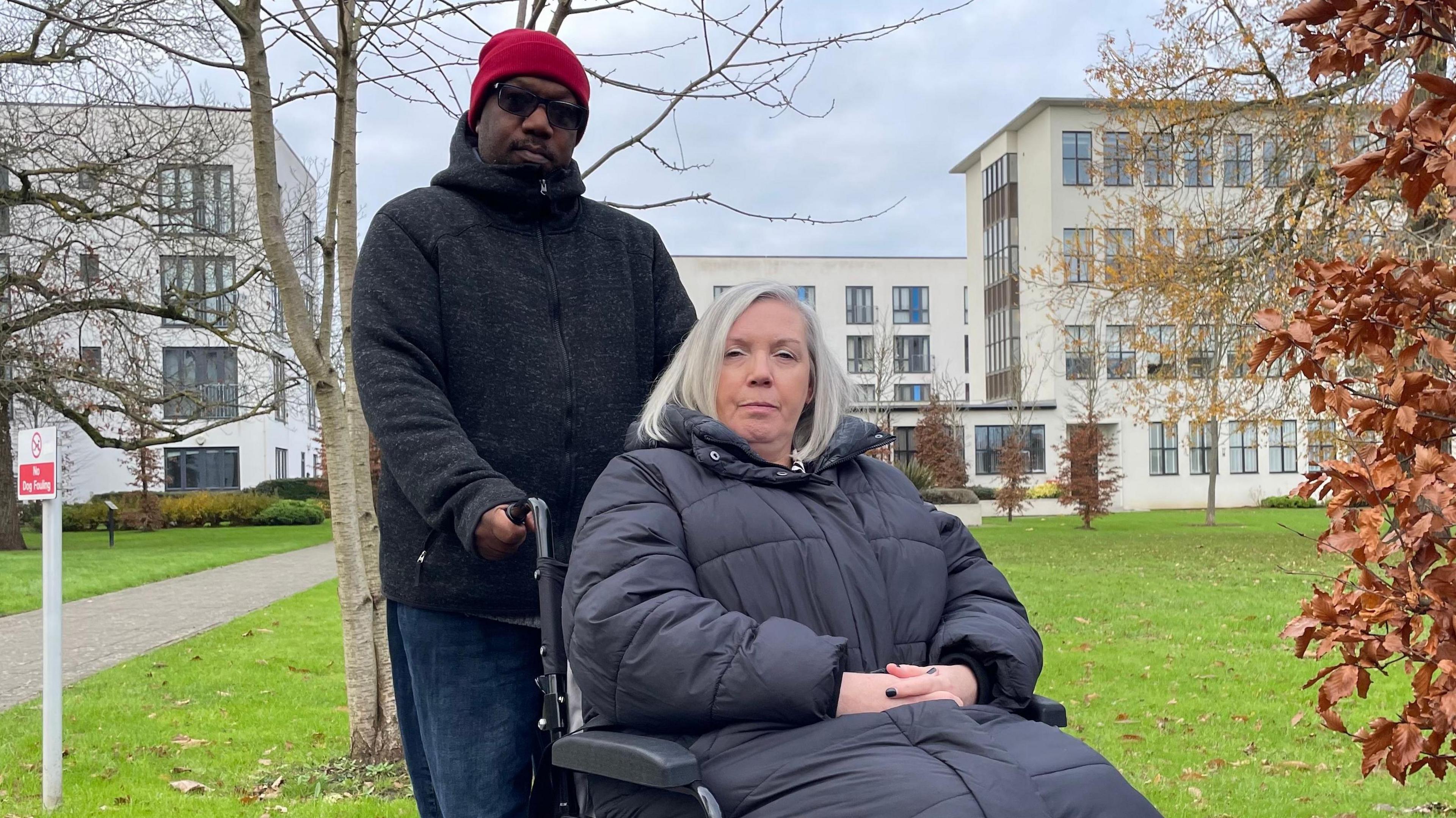 Karen James, seated in a wheelchair and looking direct to camera, with her husband Mansfield holding the chair's handles. They are outside and Karen is wearing a thick black coat that ends below her knees. She has silvery hair cut into a bob. Mansfield is standing and wearing a red bobble hat, dark glasses, a dark fleece and blue jeans. They are on the edge of a grassed area, with white art deco style buildings and some trees in the background. 