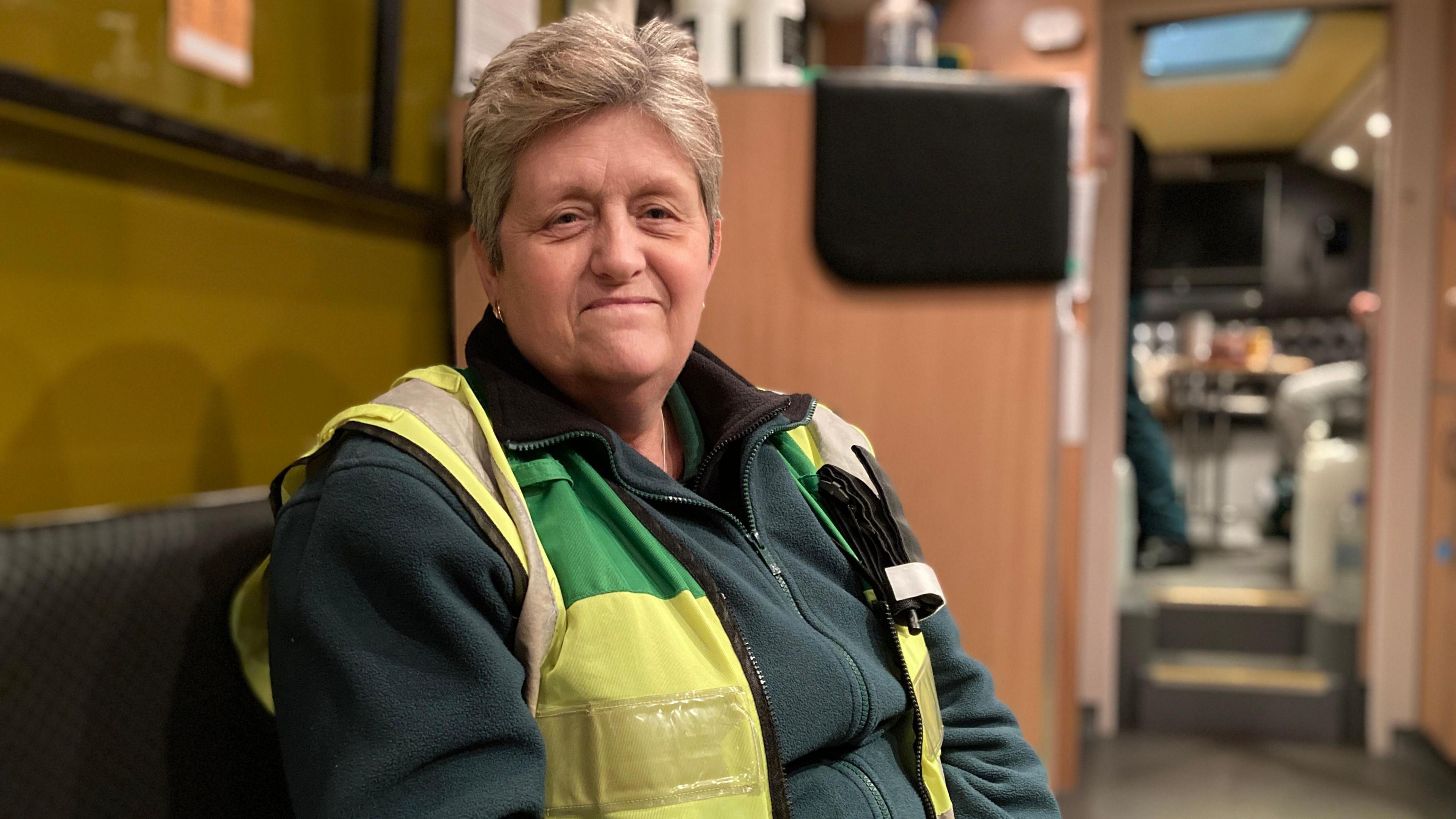 Belinda Buxton sitting on the SOS Bus. She is wearing a green jacket and yellow high-visibility vest.