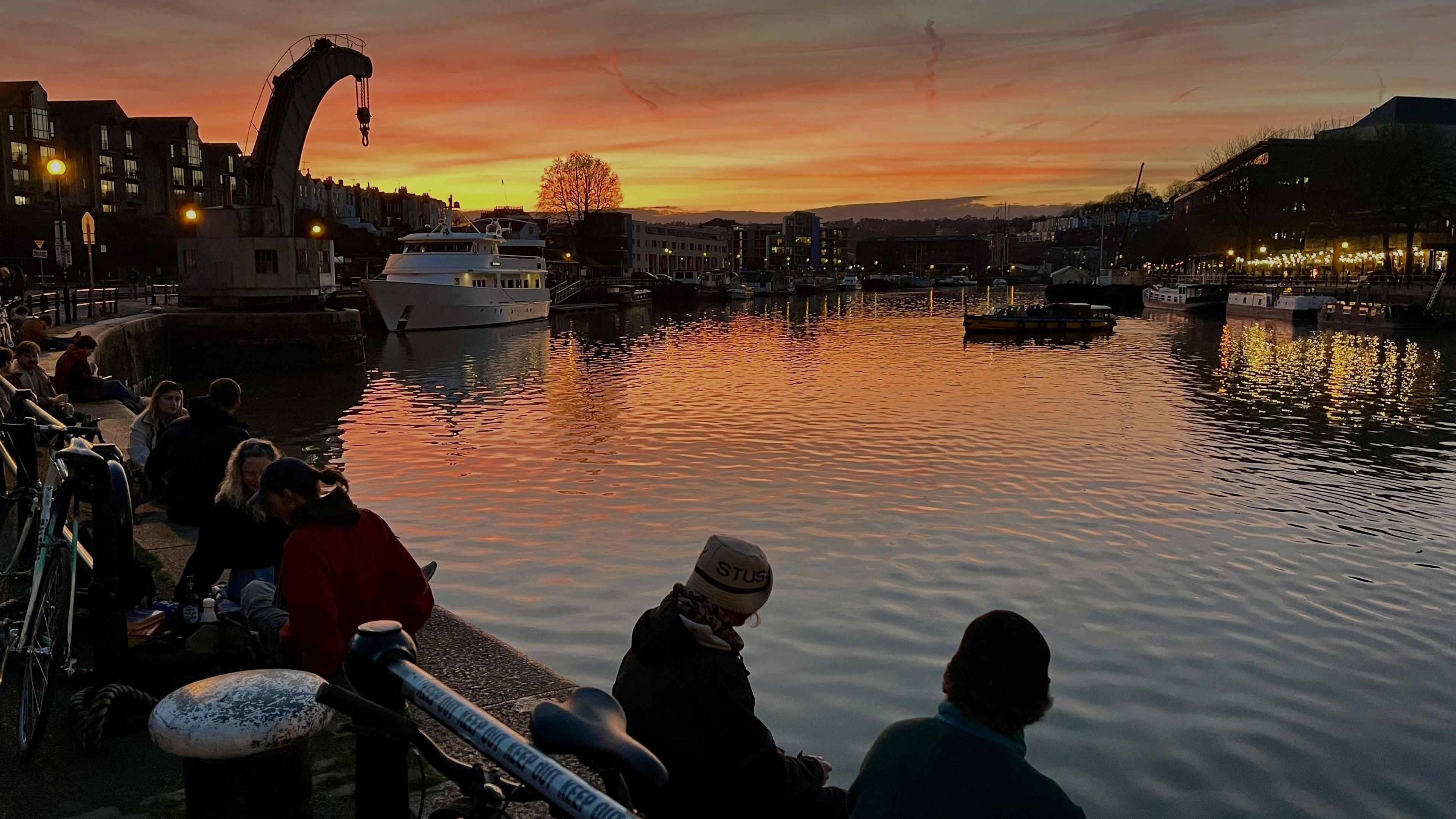 A group of people sit on the edge of Bristol Harbour facing the north side as the sun goes down, with the sky tinged with orange