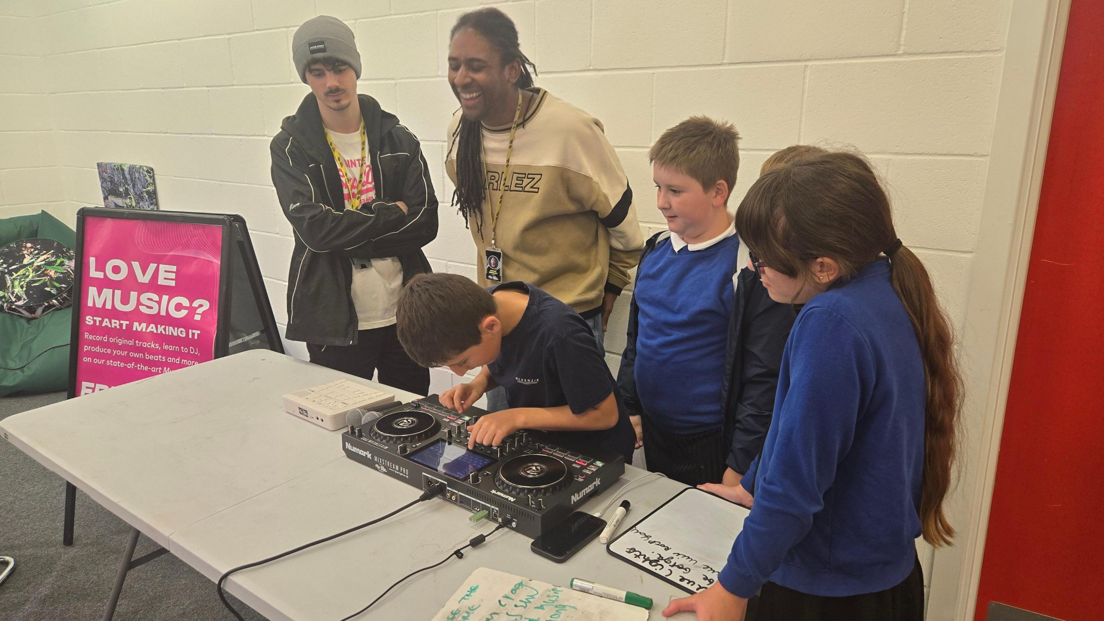 A group of four children with two adults gathered around decks learning how to DJ. They are all laughing and smiling. They're doing the session in a hall. There is a pink board next to the table saying 'Love Music? Start Making It' on it.