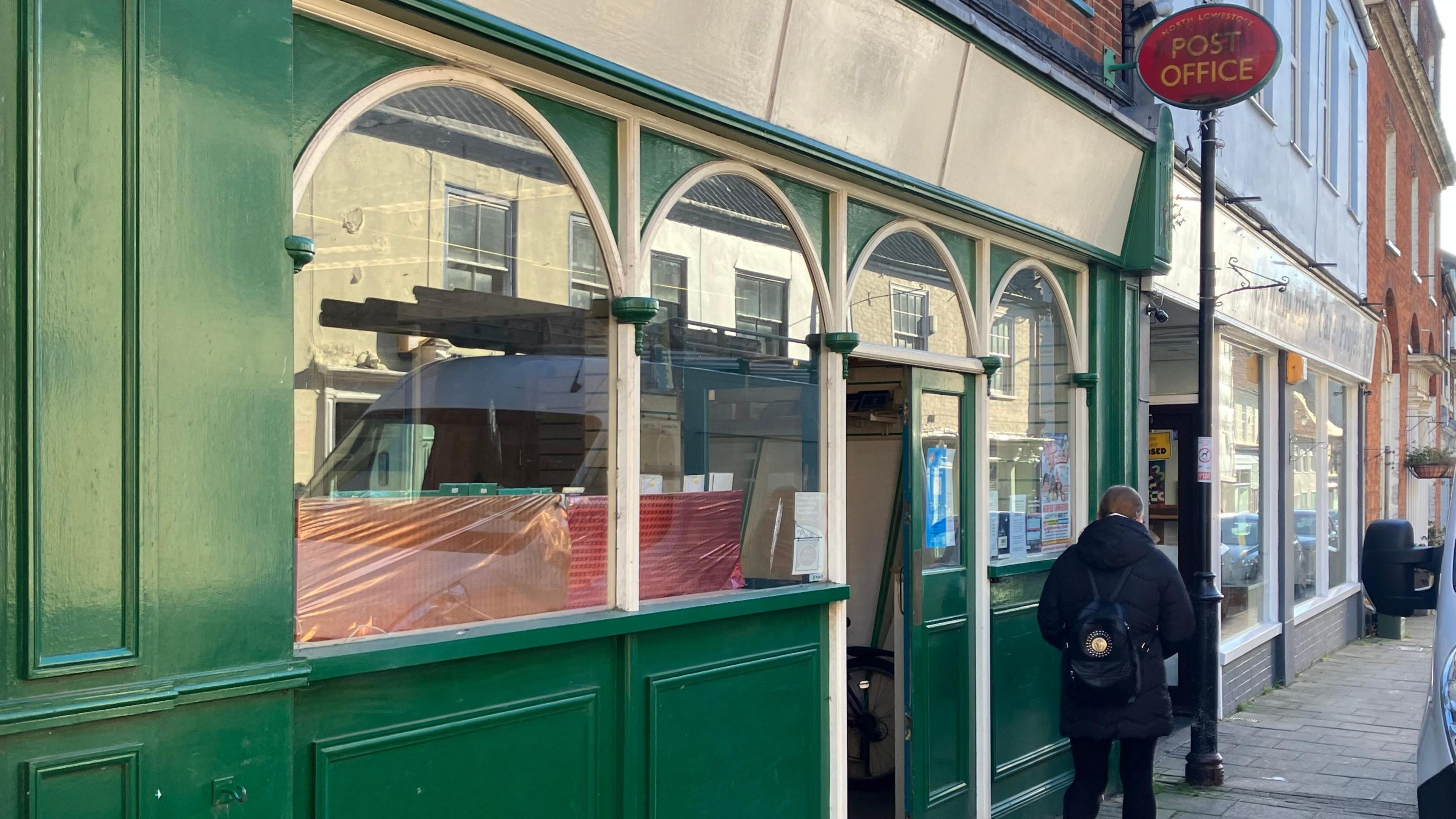 A person wearing a dark coat is walking past the green and white wooden facade of the current Post Office 