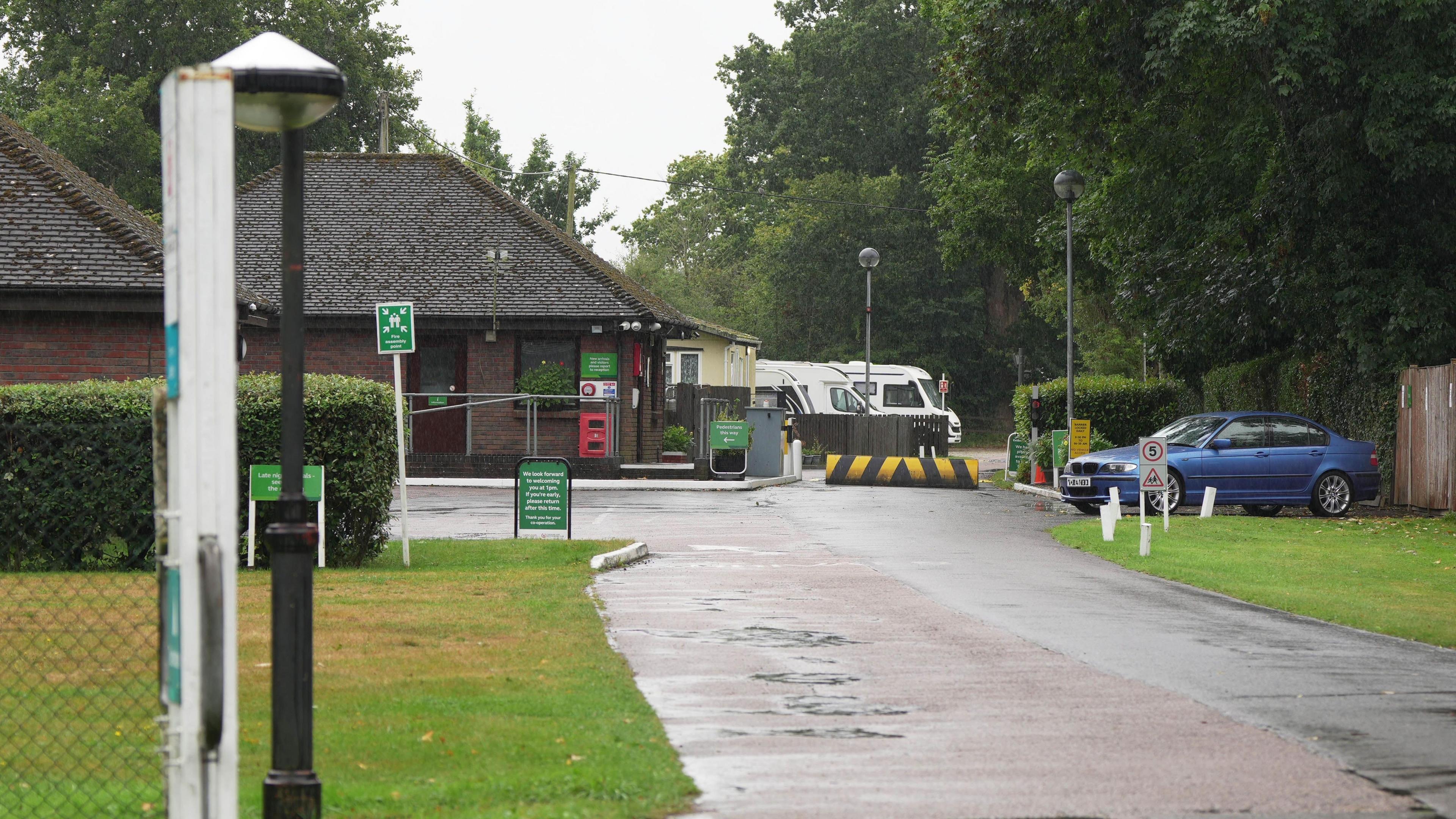 The entrance to the Gatwick Caravan and Motorhome Club Campsite, where motorhomes are seen in the background and a low brick building as well as grass and trees