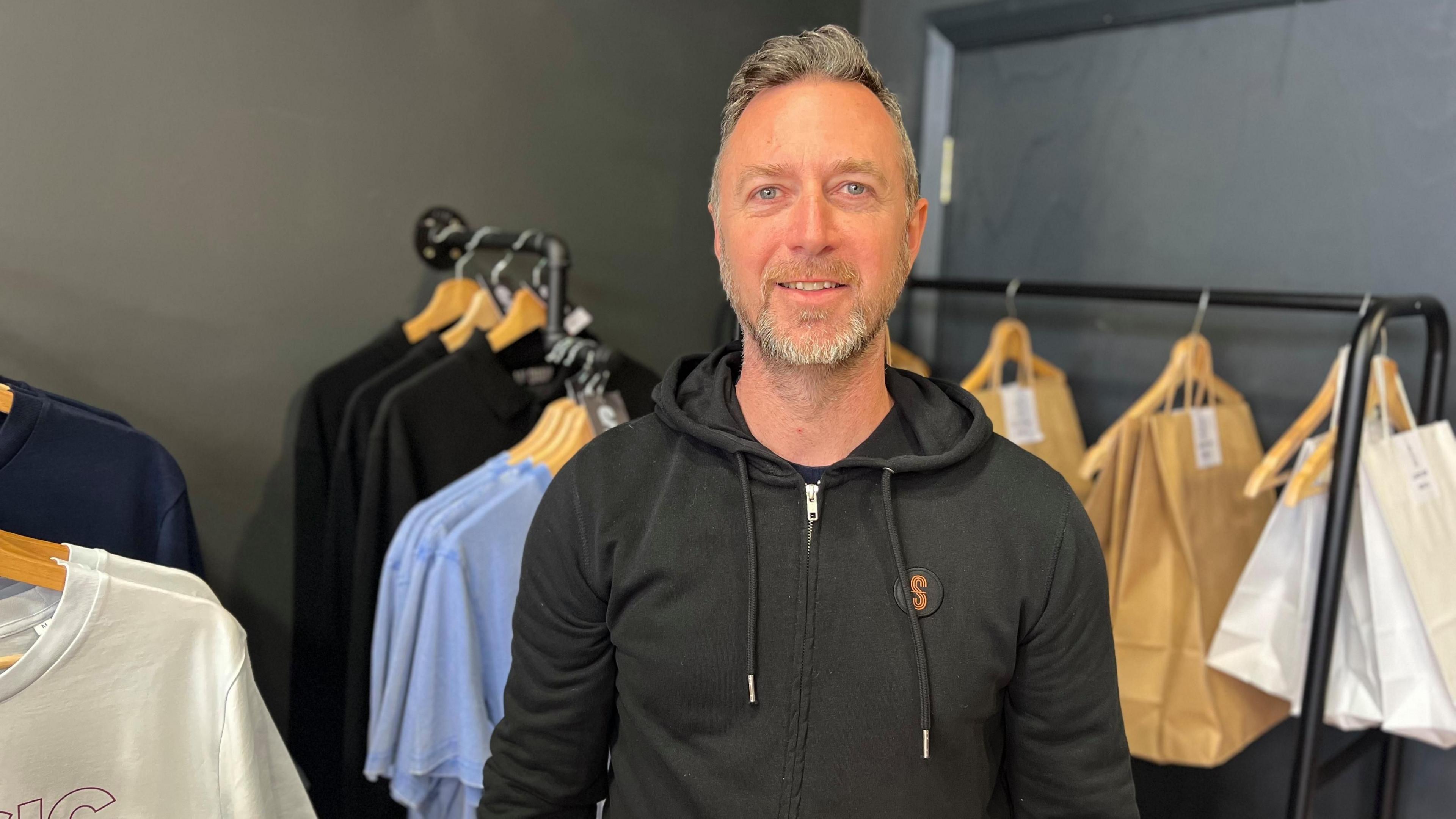 Man with grey hair and black hoodie stands infront of wall of t shirts 