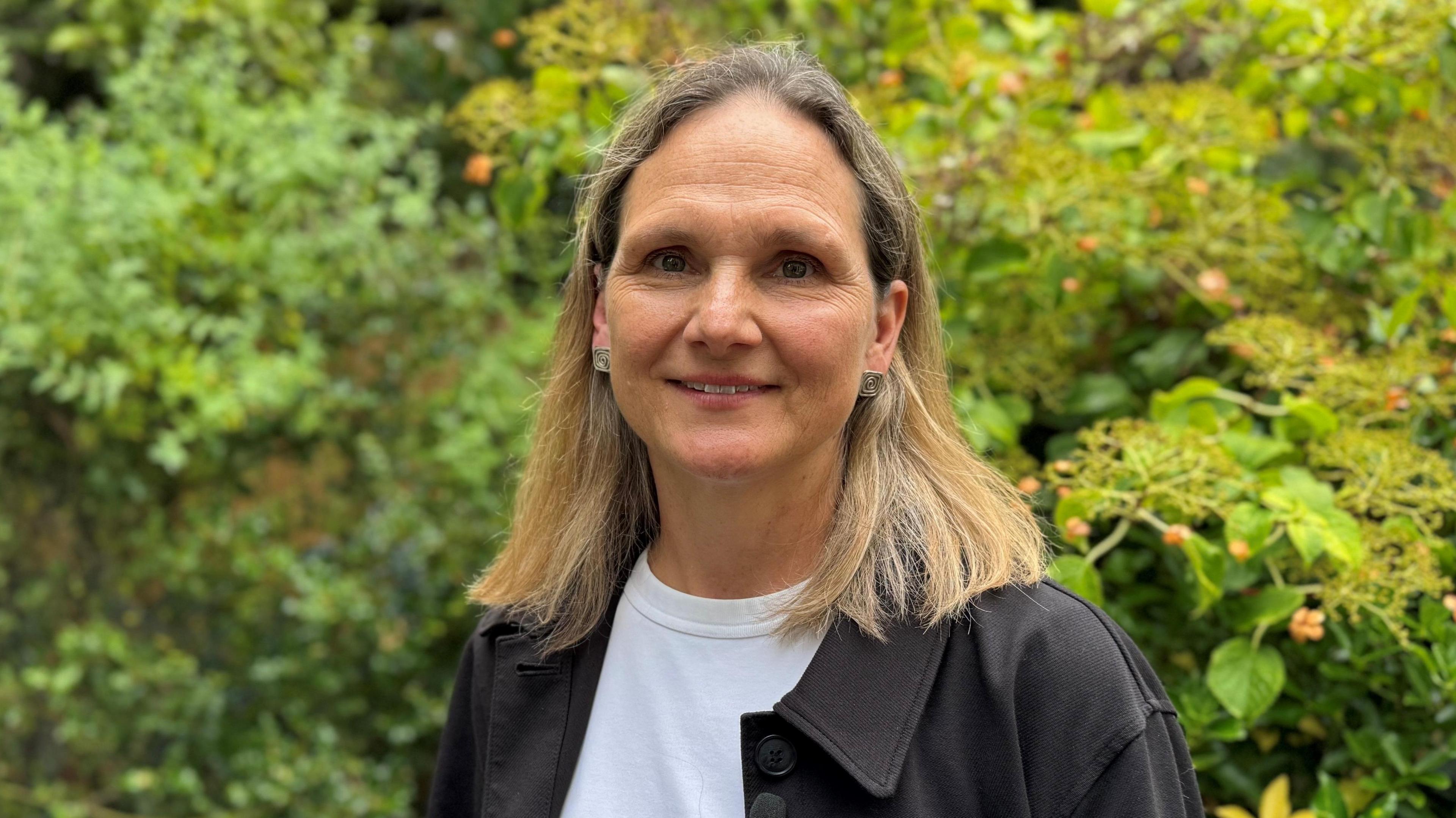 Melissa Turner pictured in her garden wearing a white top and black jacket. she has shoulder length blonde hair and is 54.
