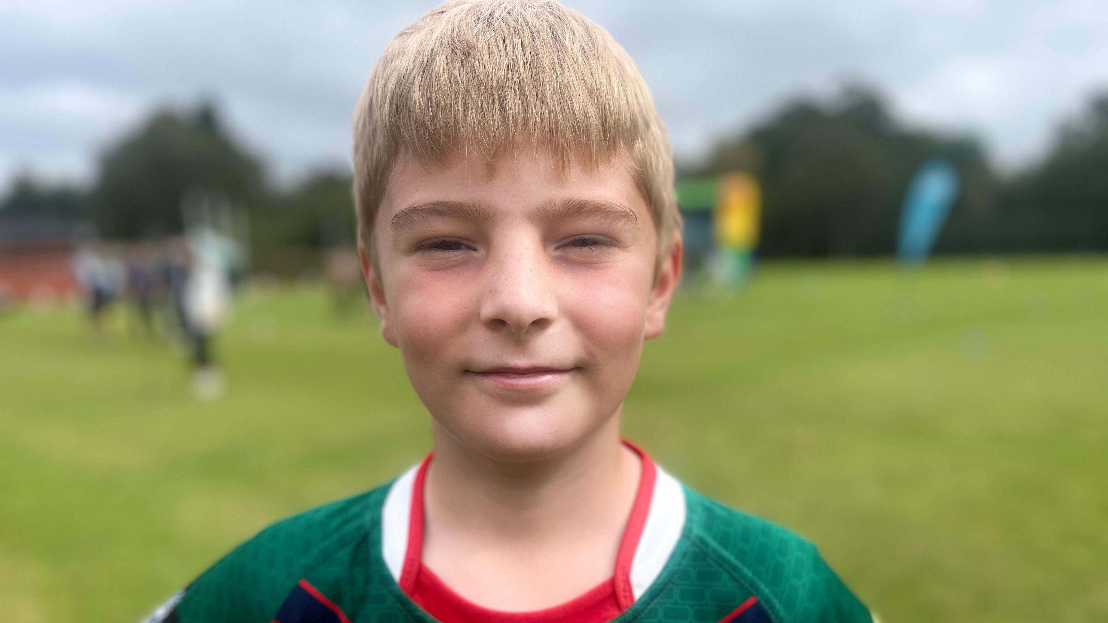 A blonde boy with a fringe stares into the camera. he has a green sports top on but it cant be seen - just the collar.