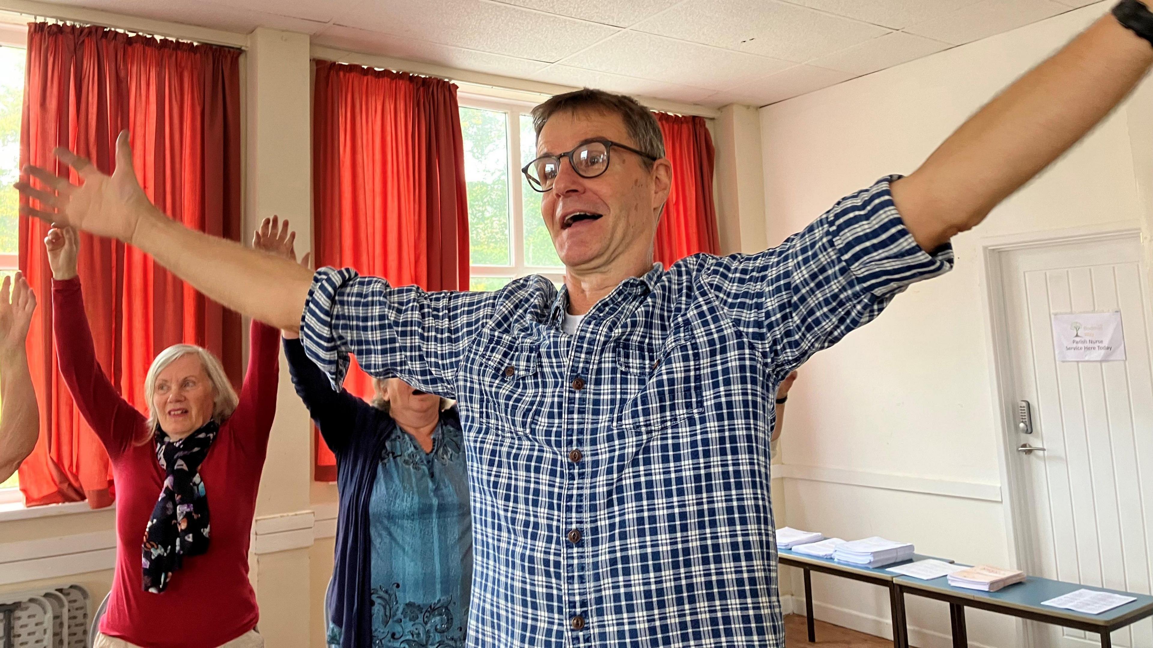 Martin Palmer wears a checked shirt and has short dark hair glasses. He has arms stretched up in the air. Two older ladies behind him are doing the same.