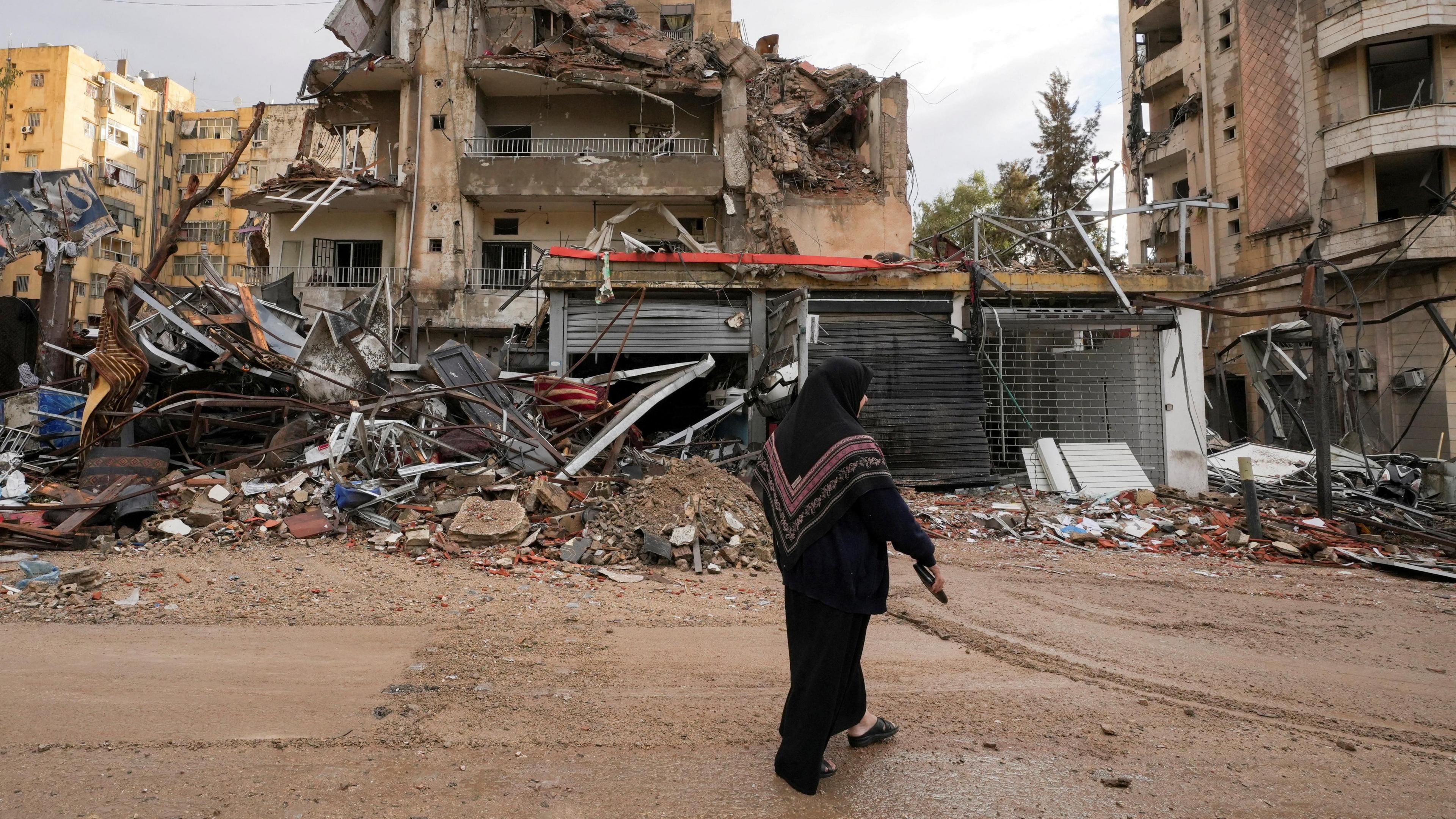 A woman dressed in a black headscarf and dark clothing walks past the wreckage of heavily damaged buildings in Beirut’s southern suburbs, Lebanon. The scene shows extensive destruction, with collapsed walls, twisted metal, and debris scattered across the ground.