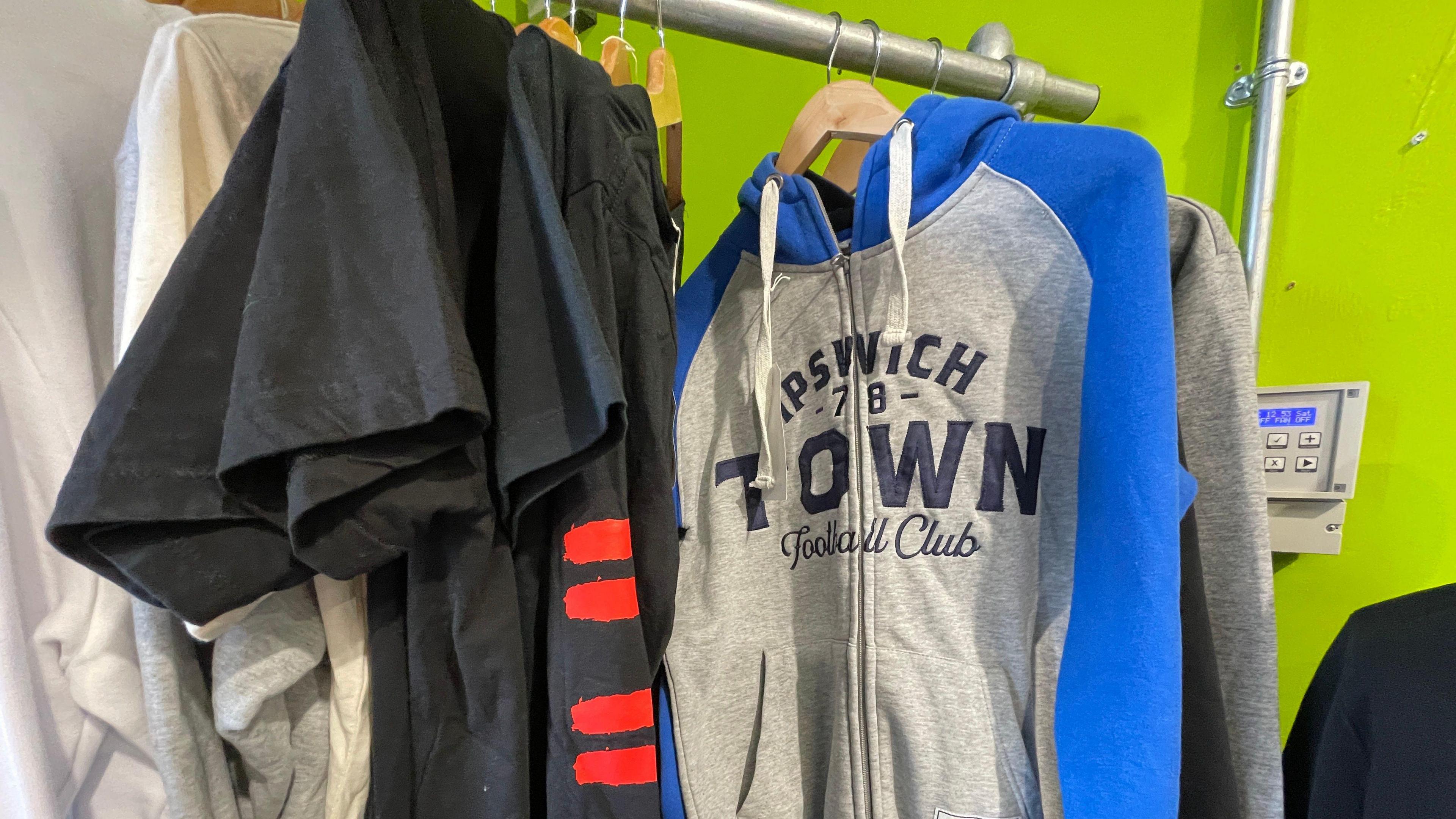 A blue and grey Ipswich Town Football Club hoody hanging on a silver rail alongside dark t-shirts