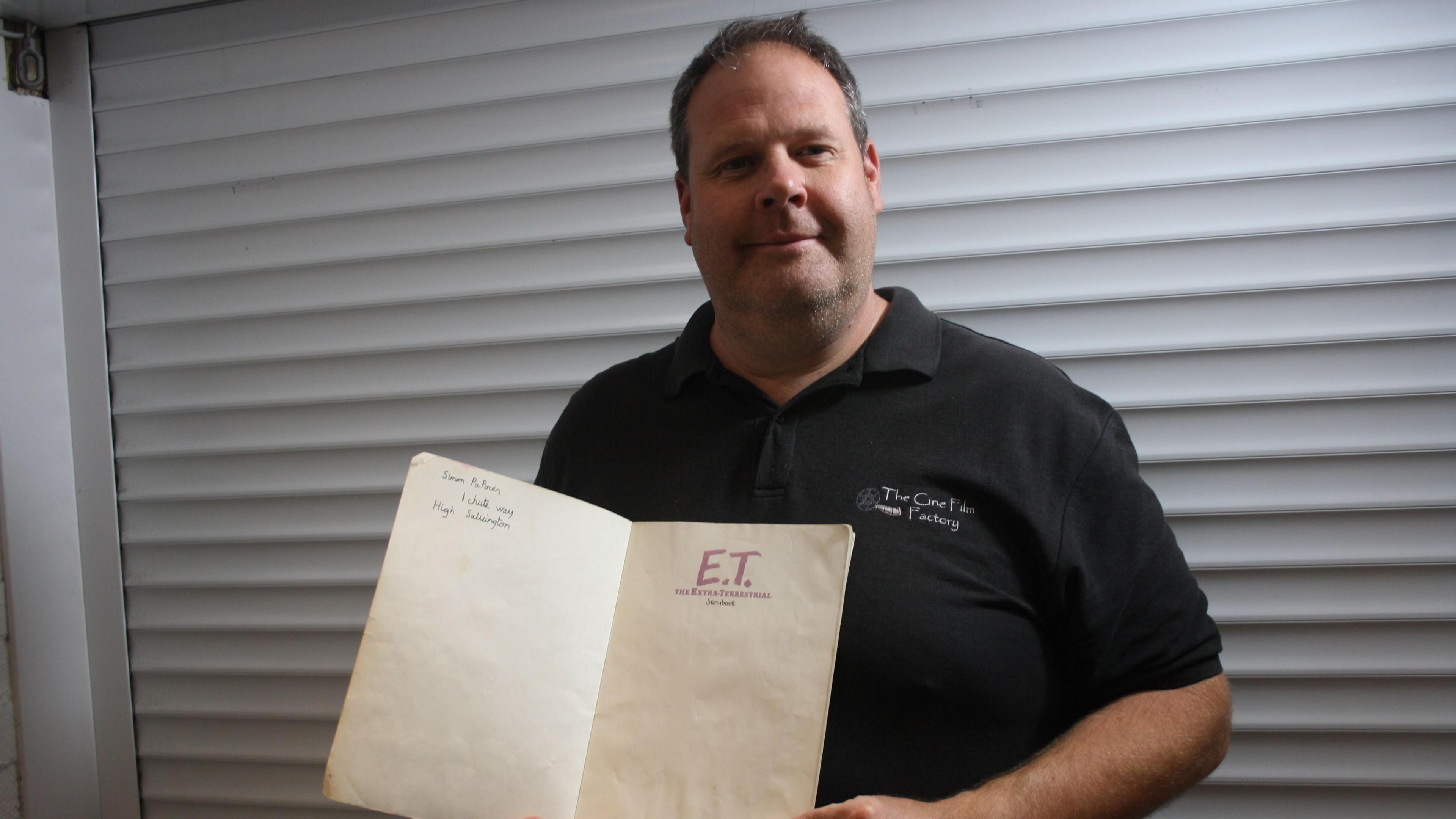 Simon Papouis wearing a black polo shirt and standing in front of white metal shutters. He is holding an open book with handwritten words on the left-hand page and the words "E.T the extra terrestrial" in red capital letters on the right-hand page