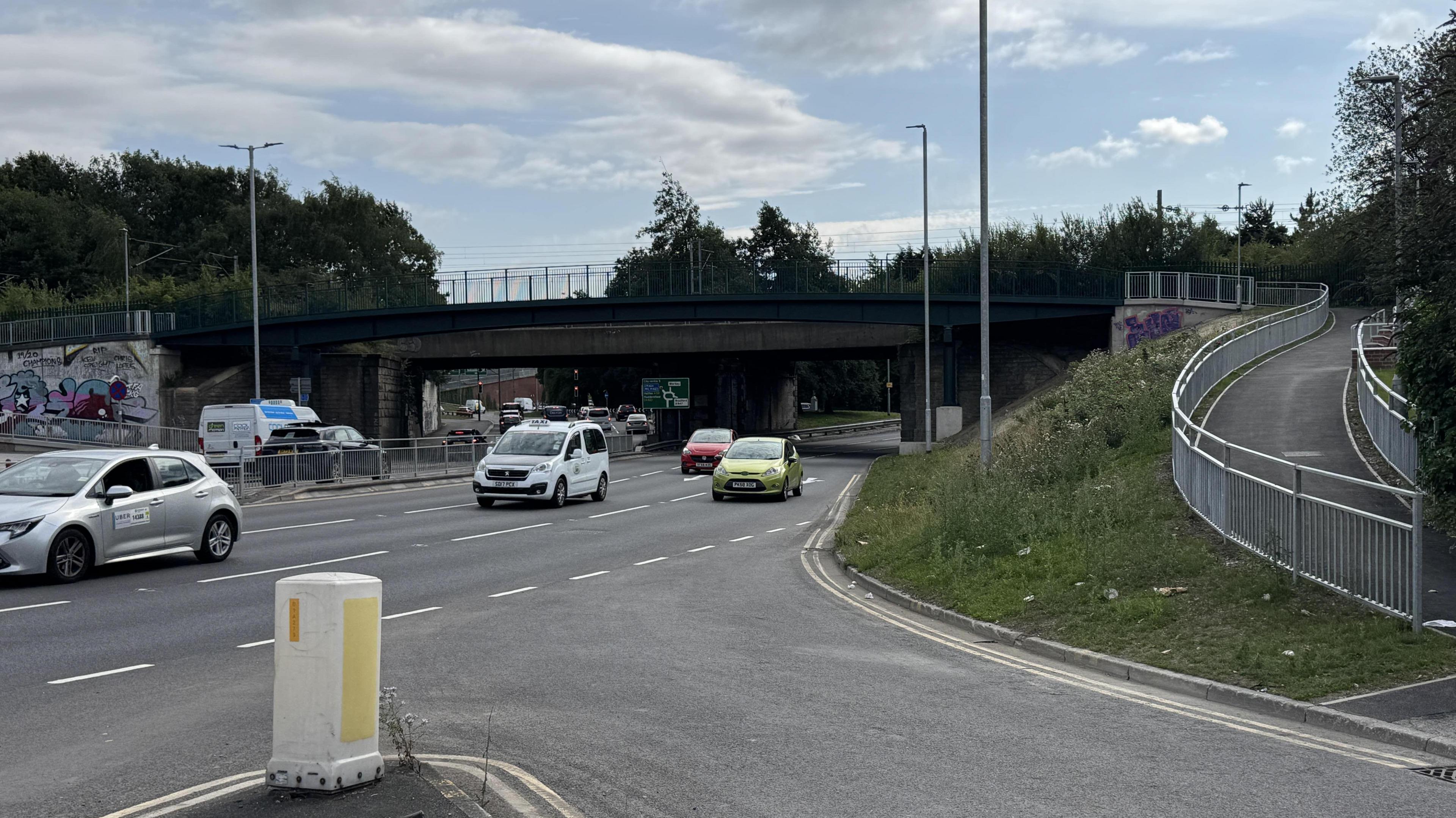 Cars go by on a three-lane road. A traffic bollard is in the foreground. To the side is a footpath which slowly escalates uphill.
