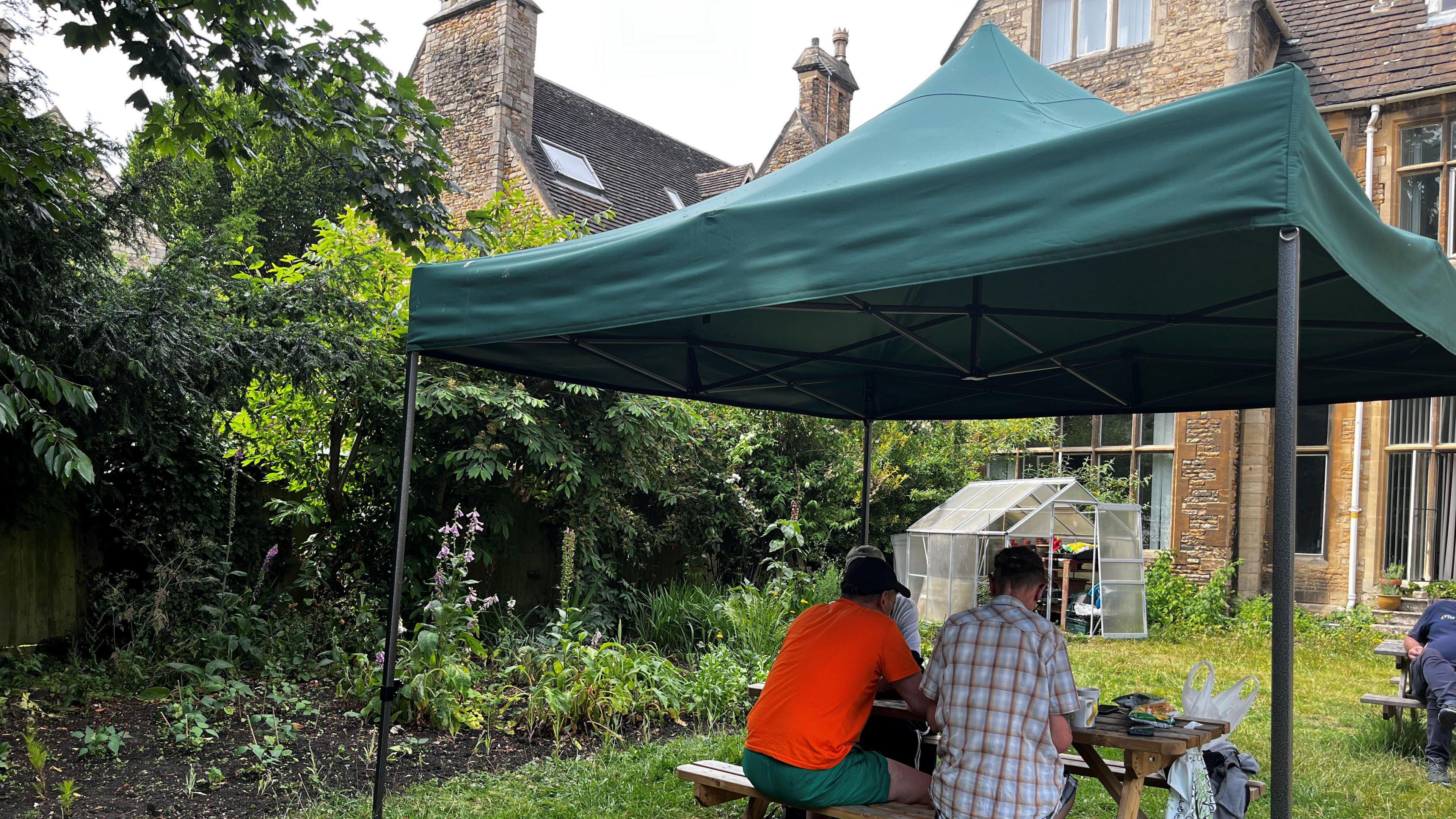 A green gazebo with two people sitting underneath it, in a garden, in the background is a stone building.