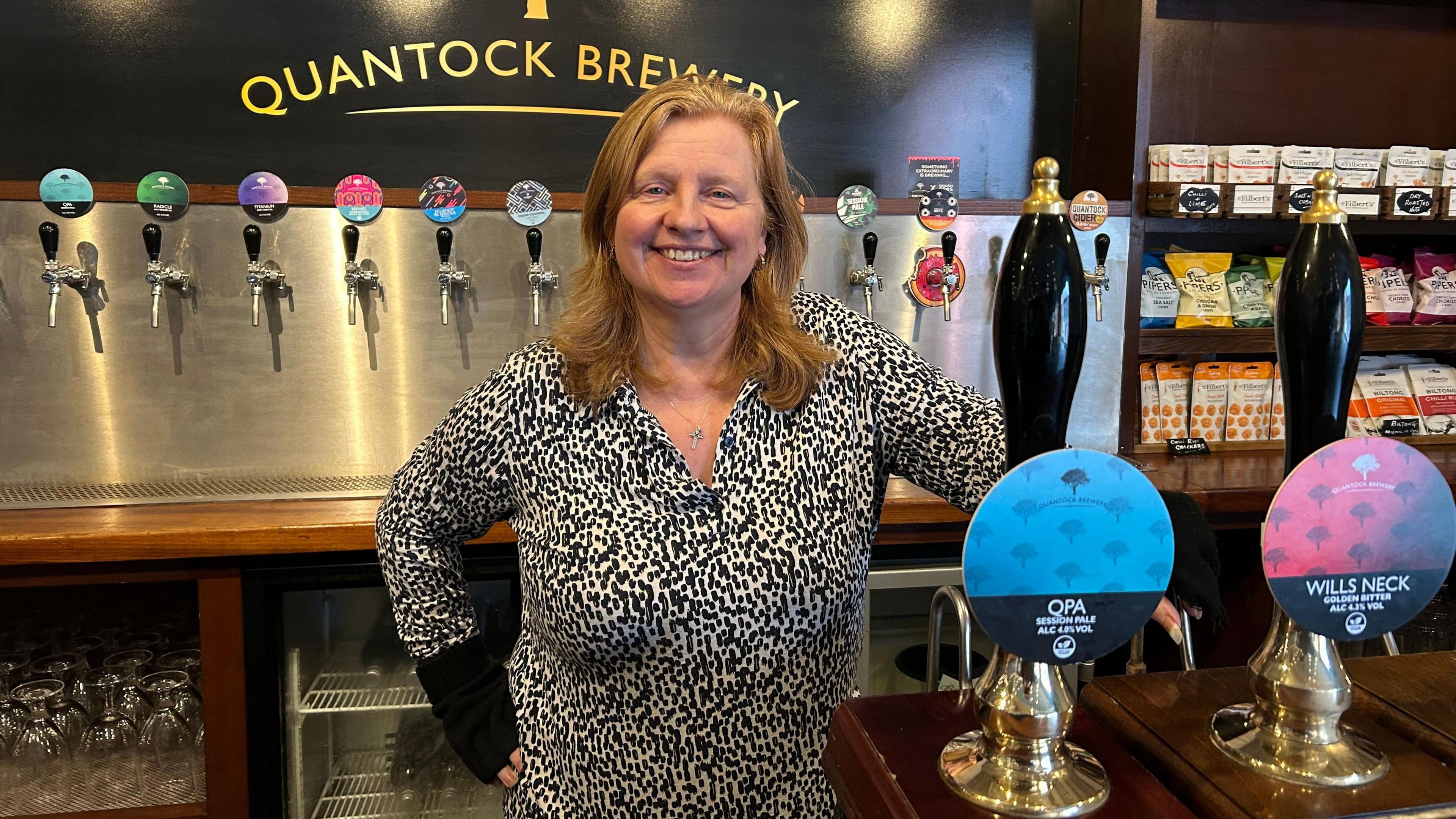 Cheryl Ford stands smiling behind the bar at Quantock Brewery. She has shoulder length dark blonde hair and is wearing a black and white spotted blouse. She has one hand on her hip and the other resting behind the bar.