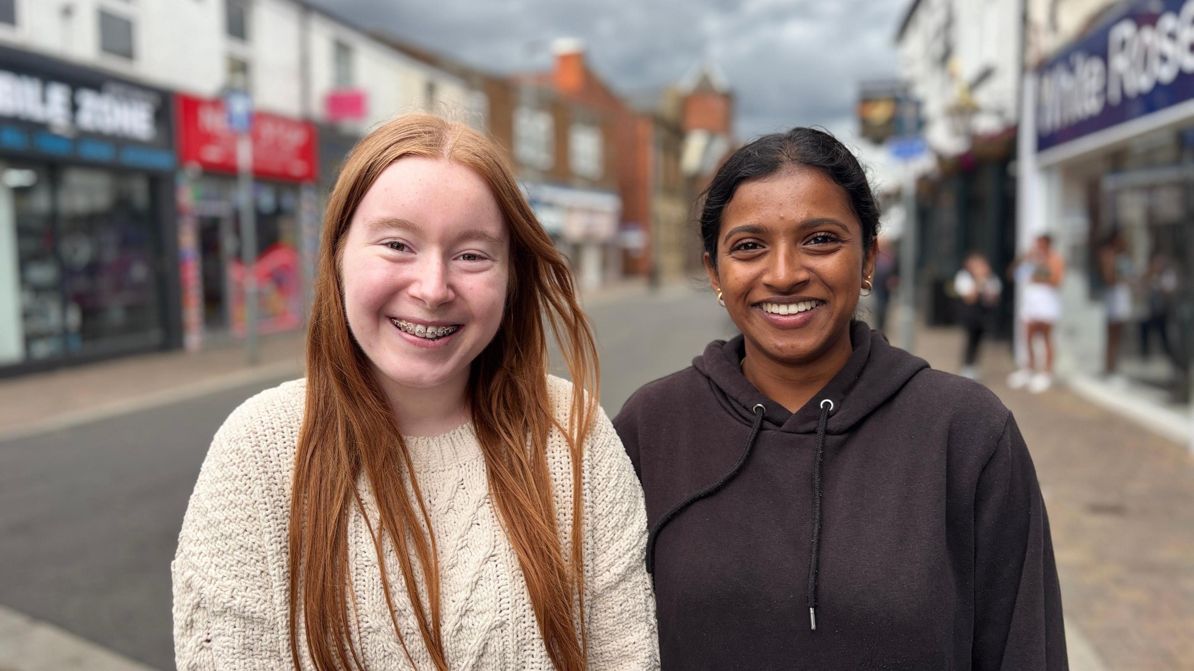 A lady wearing a white jumper with long red hair is on the left of the picture. To her right is a lady wearing a black hoodie with earrings. Behind them is a street scene