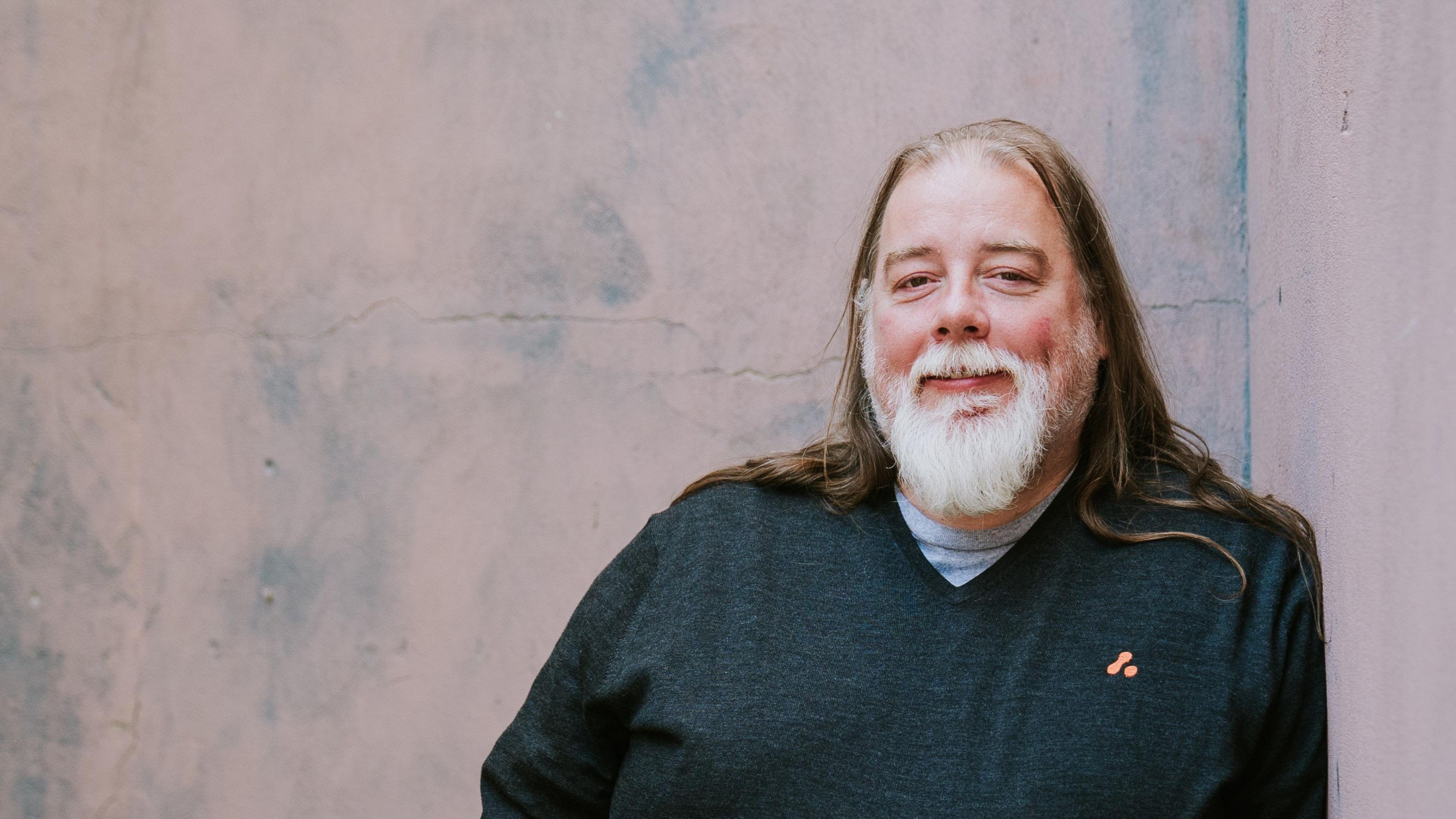 Long-bearded Simon Haighton-Williams, smiles while leaning against a wall.