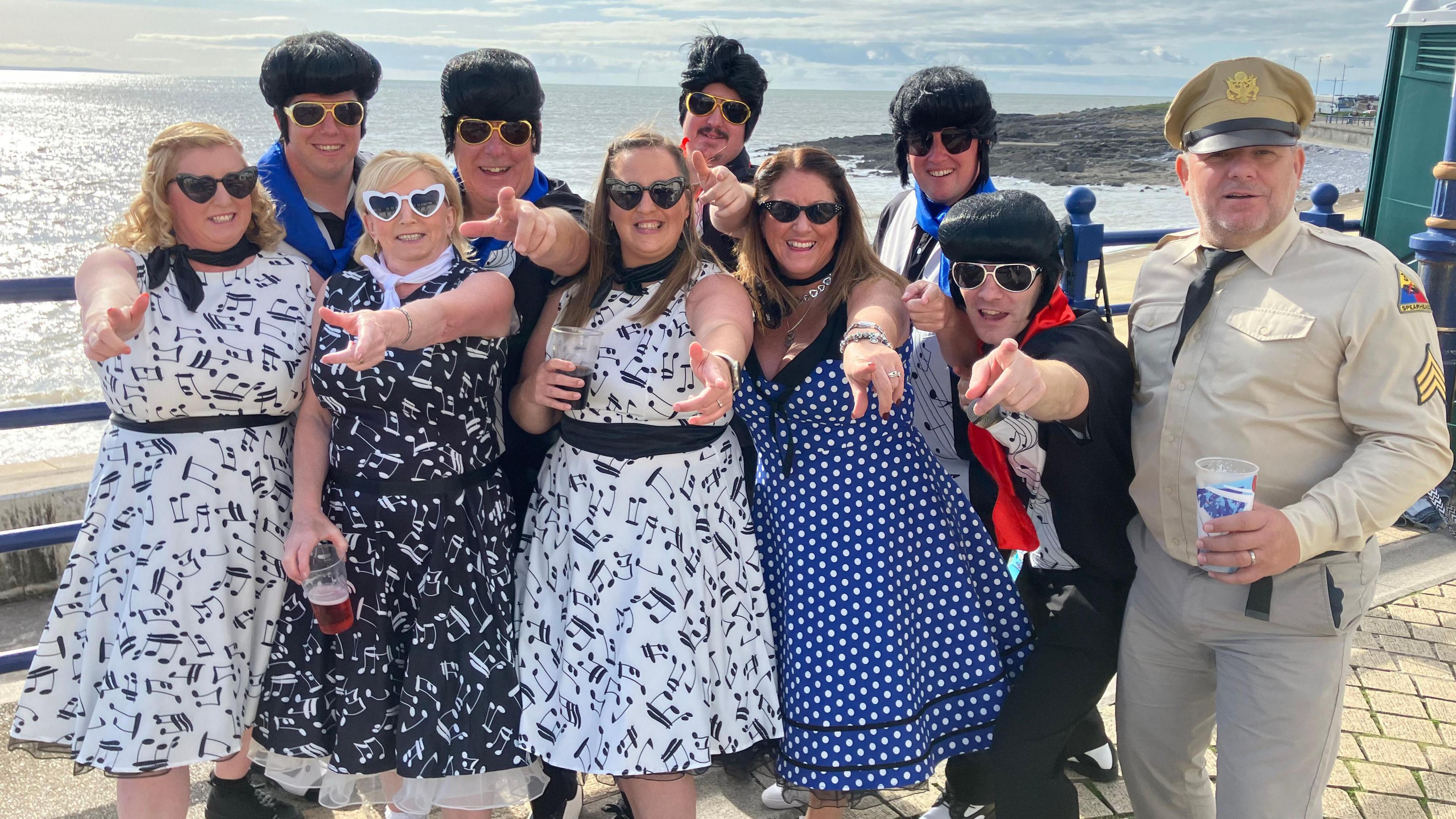 Five Elvis impersonators, a man dressed in uniform and a four women in 50s style dresses