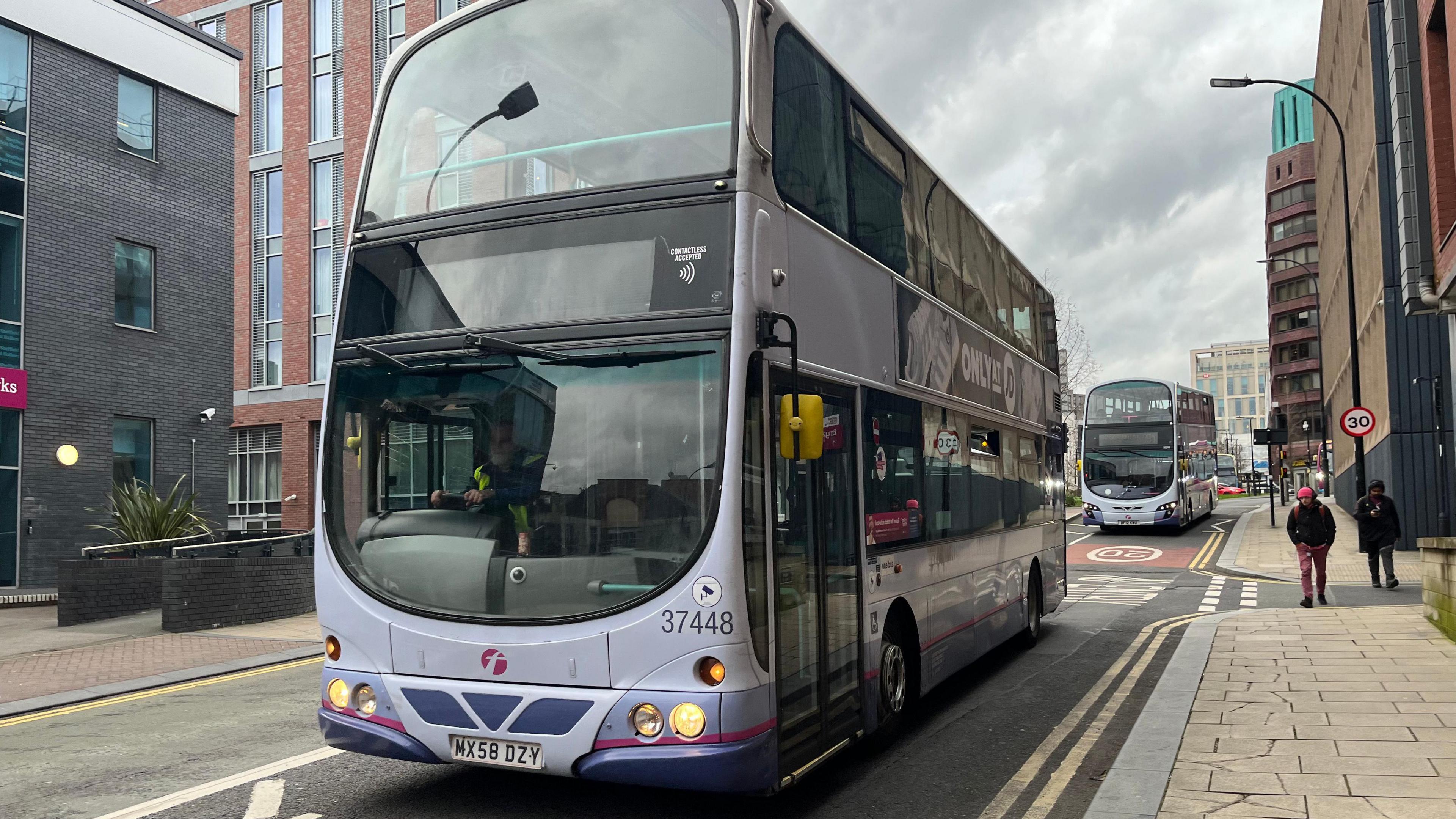 A First Bus stopped on a city centre road. The bus is coloured various shades of purple with some advertisement on its sides. People walk on the pavement next to it. 