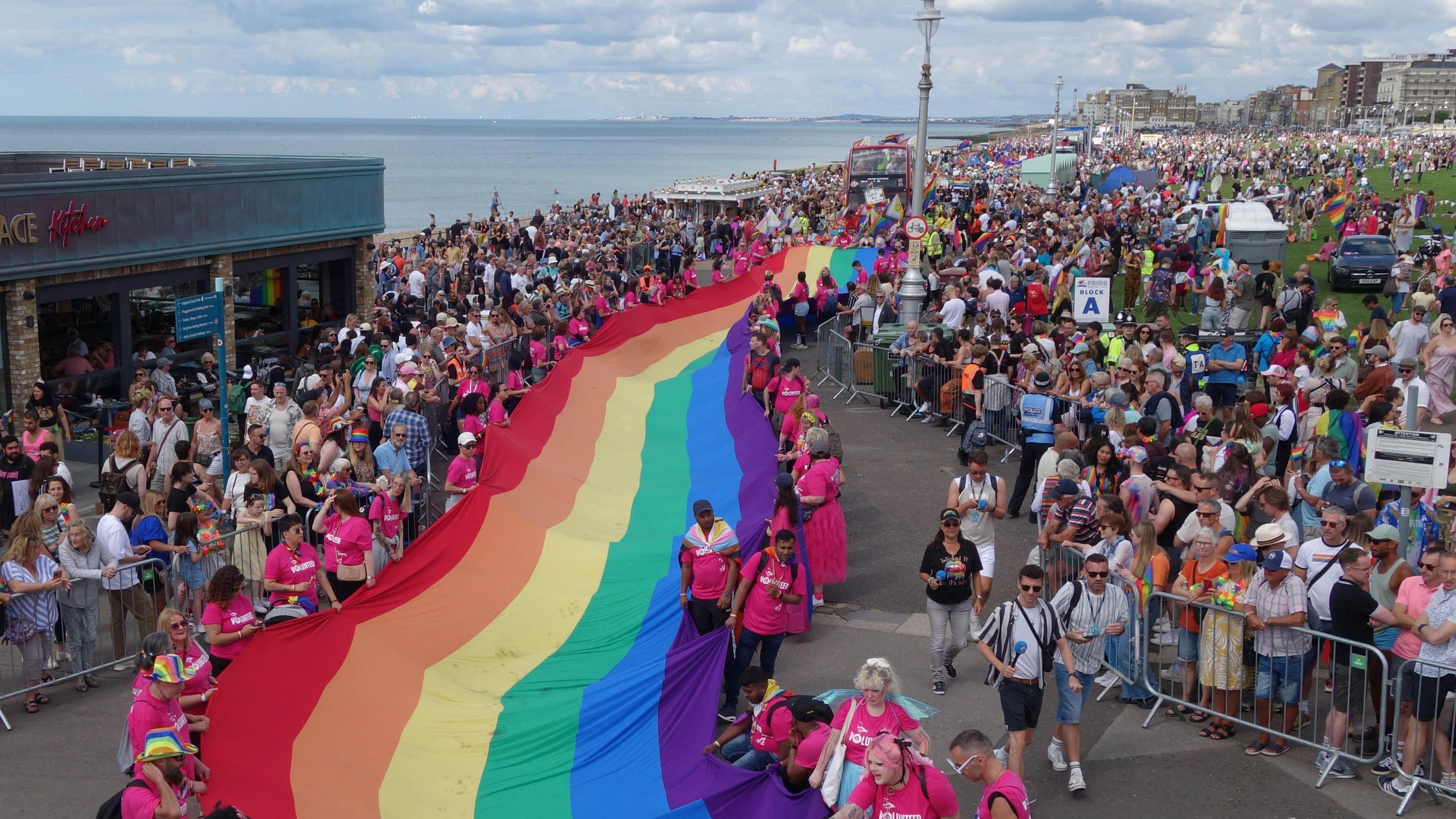 A group holding a rainbow flag during the Brighton Pride March on Saturday 2 August, 2025. Groups of onlookers can be seen either side of the flag.