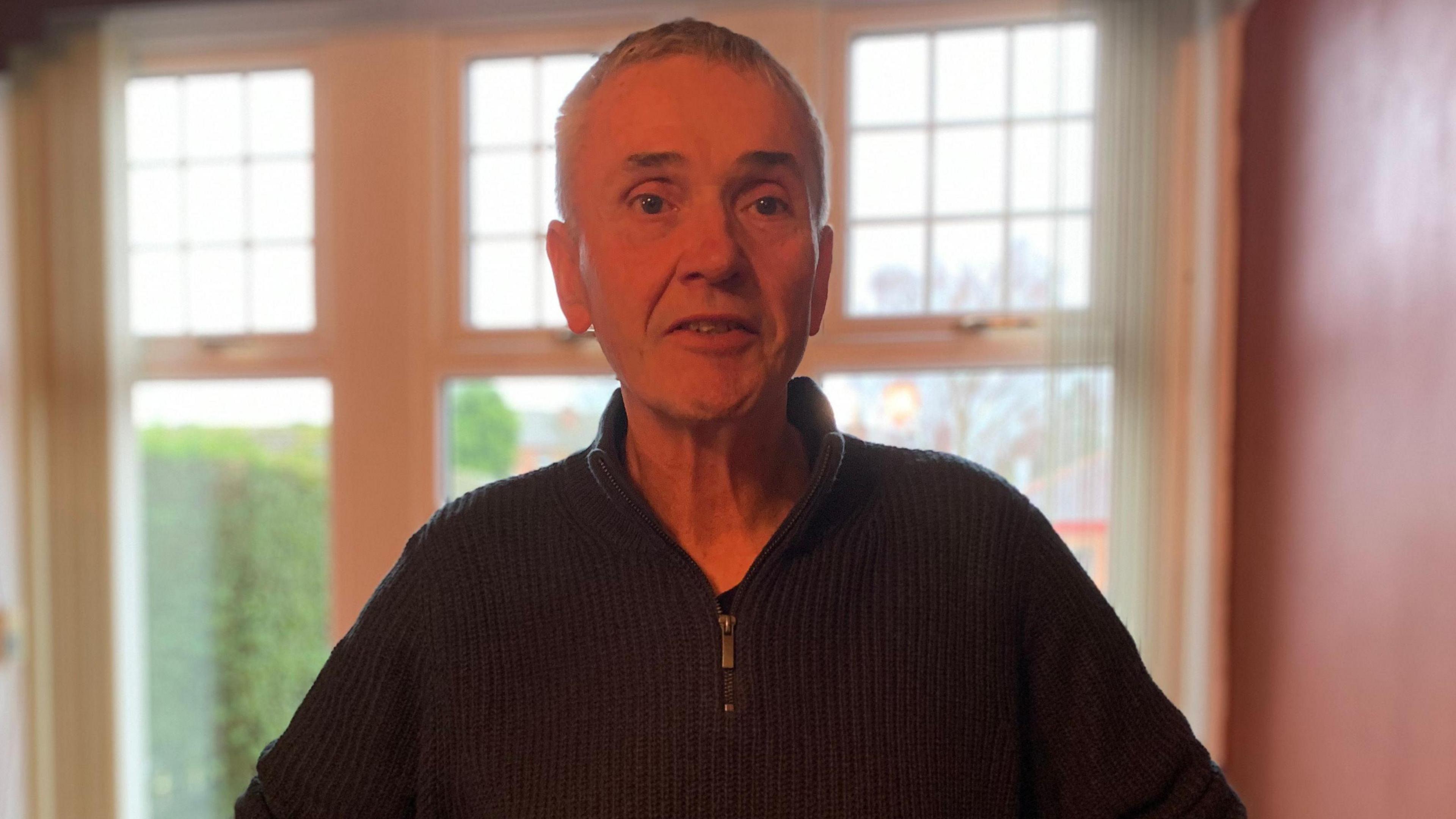 A man with grey hair and a zip-top blue jumper stands in front of a window in his living room