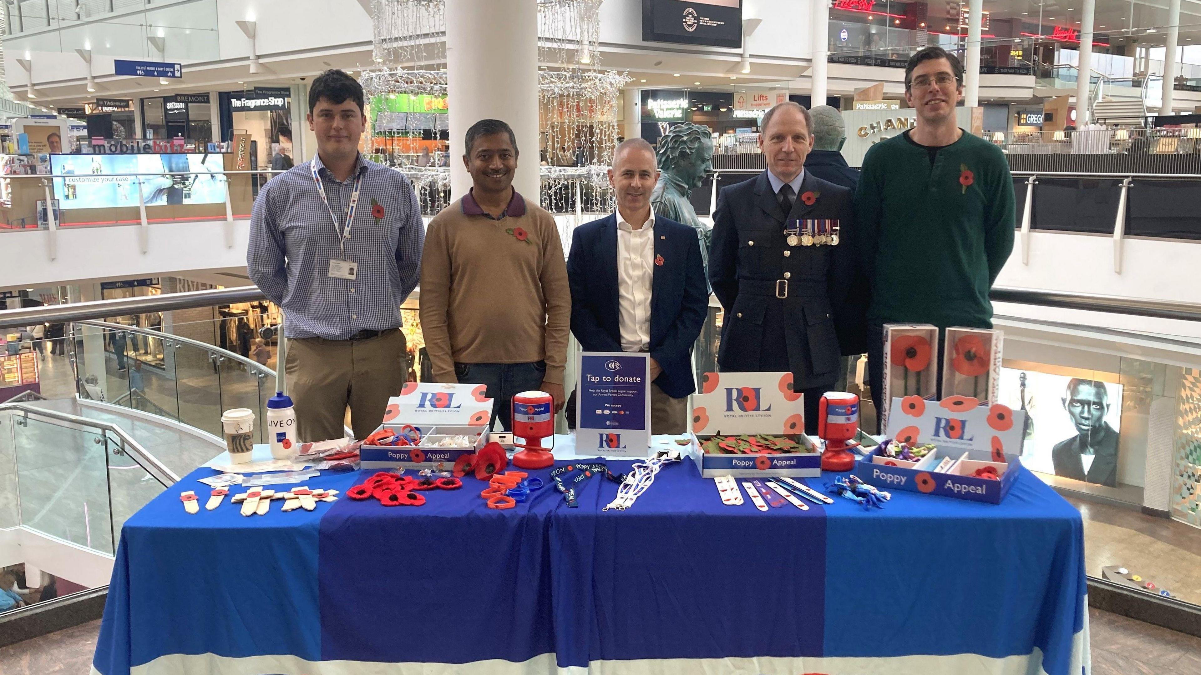 A group of five men, one of them in a military uniform with medals on it, stand beside a table with Royal British legion branding on it. On the table are poppies for sale along with collection boxes and other merchandise