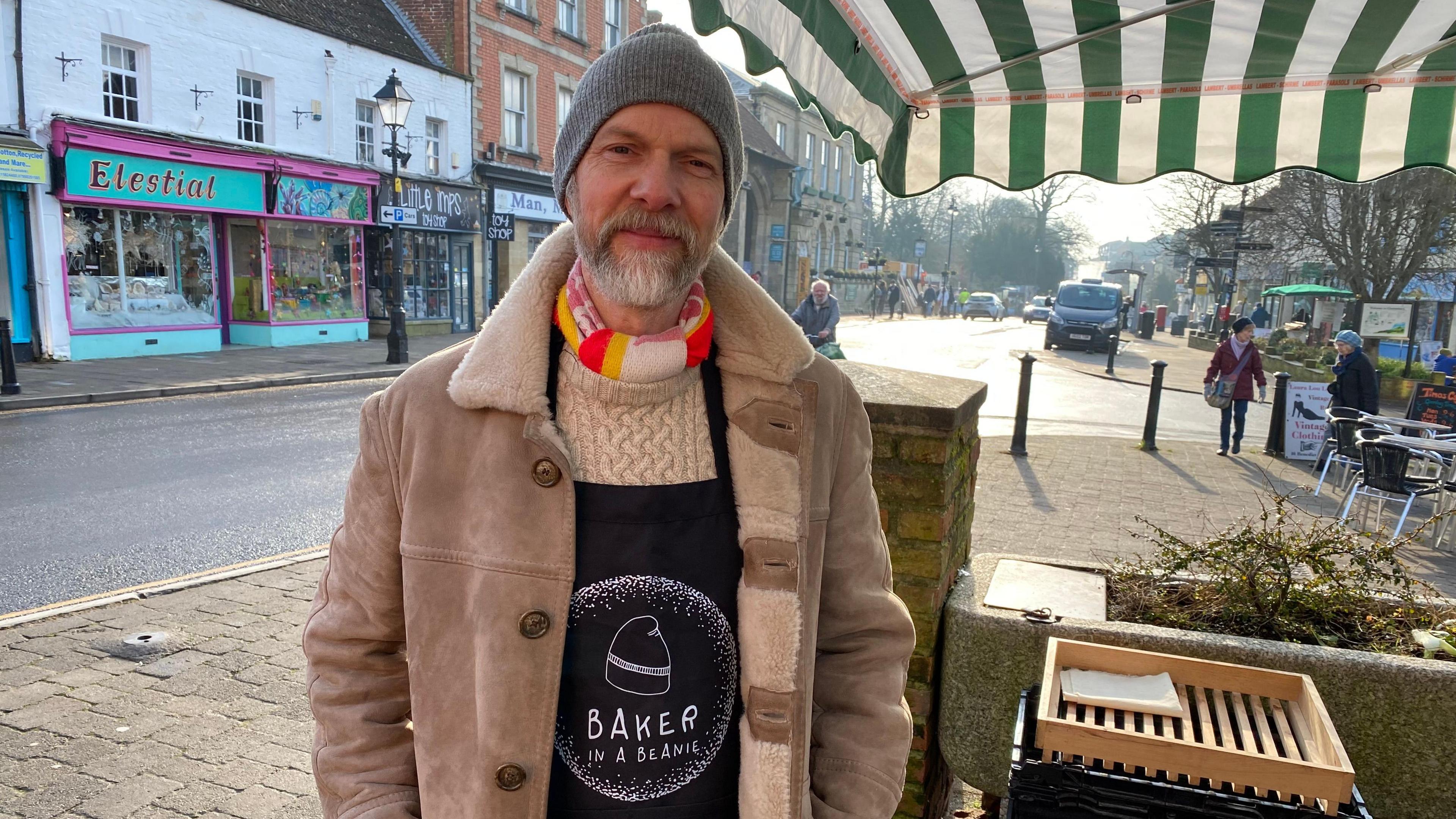 Ian Roberts is standing alongside his stall, Baker in a Beanie, in Market Cross.  He is wearing a big coat, scarf, hat and apron with Baker in a Beanie written on it. 