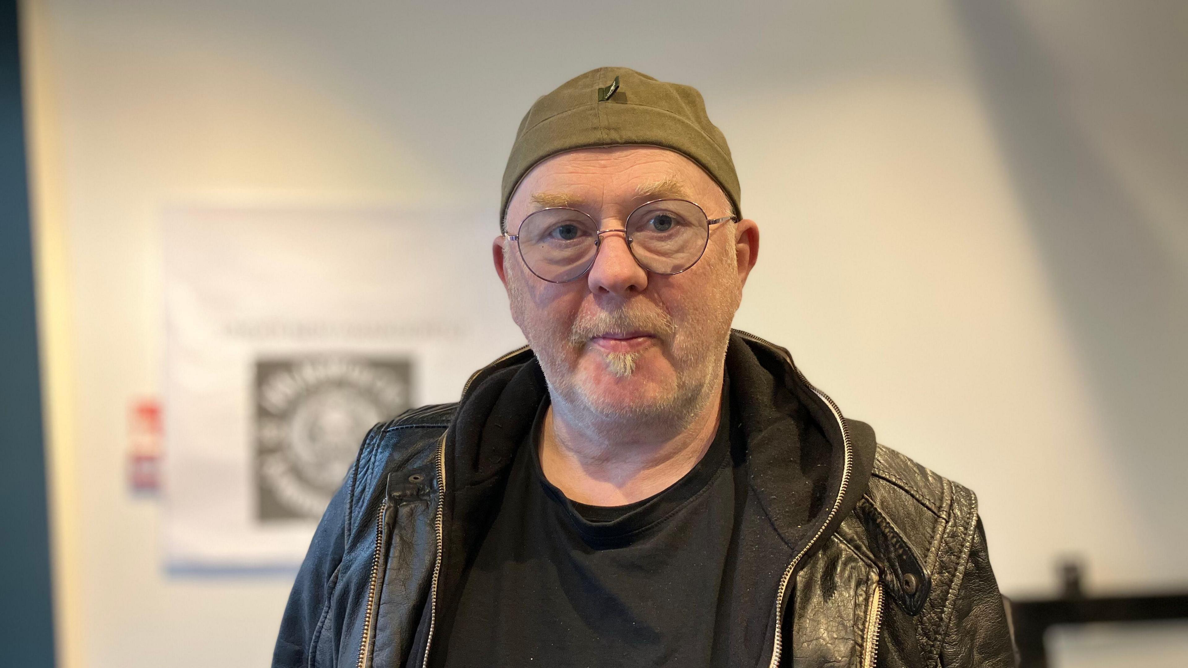 A man with a grey moustache and small goatee, wears round glasses in a thin frame. He is also wearing a black leather jacket with a black hoodie underneath, and is looking directly at the camera.