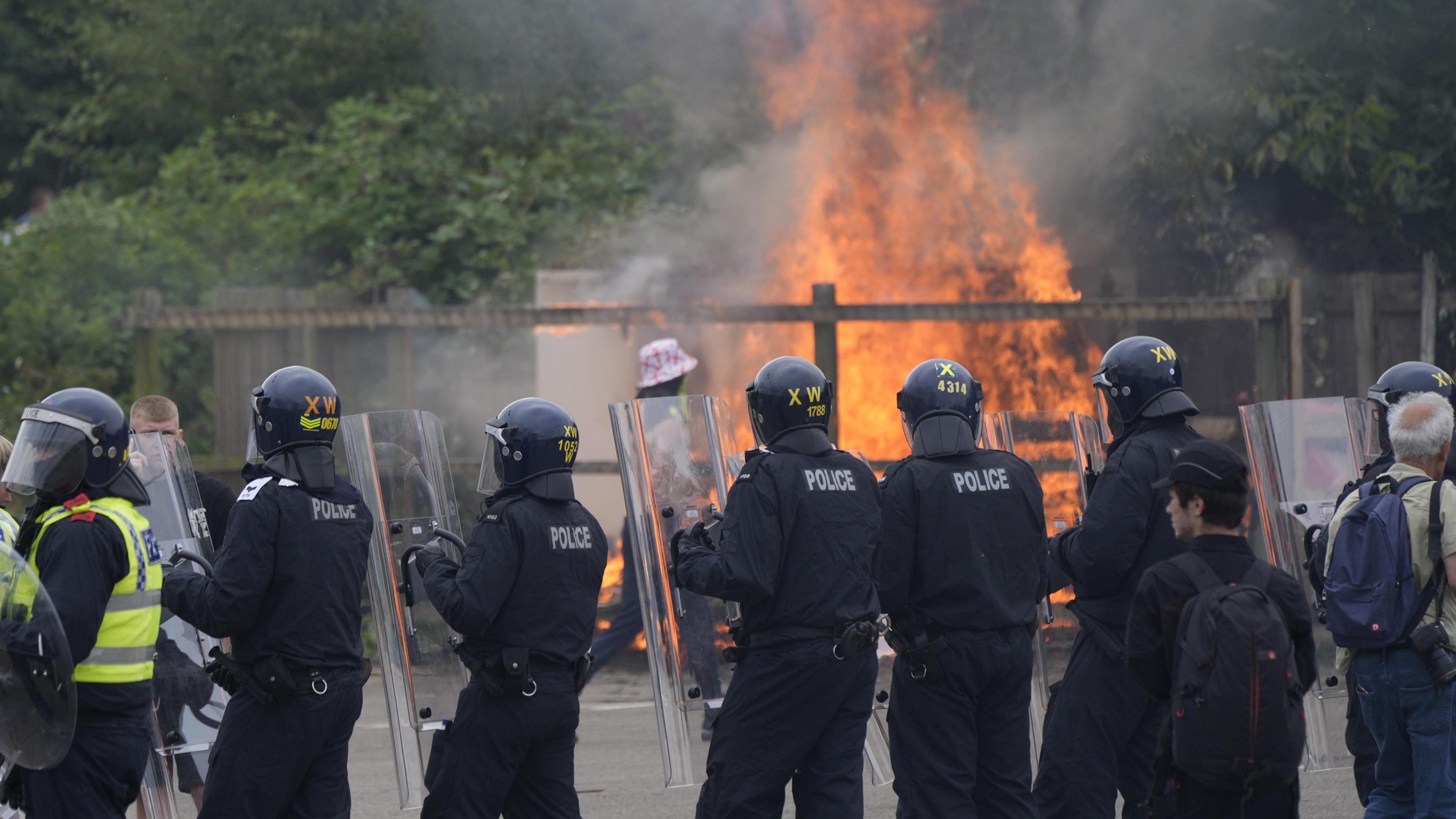A line of officers with riot shields and helmets stand with their back to the camera, facing a large fire near some trees at the Holiday Inn Express in Rotherham