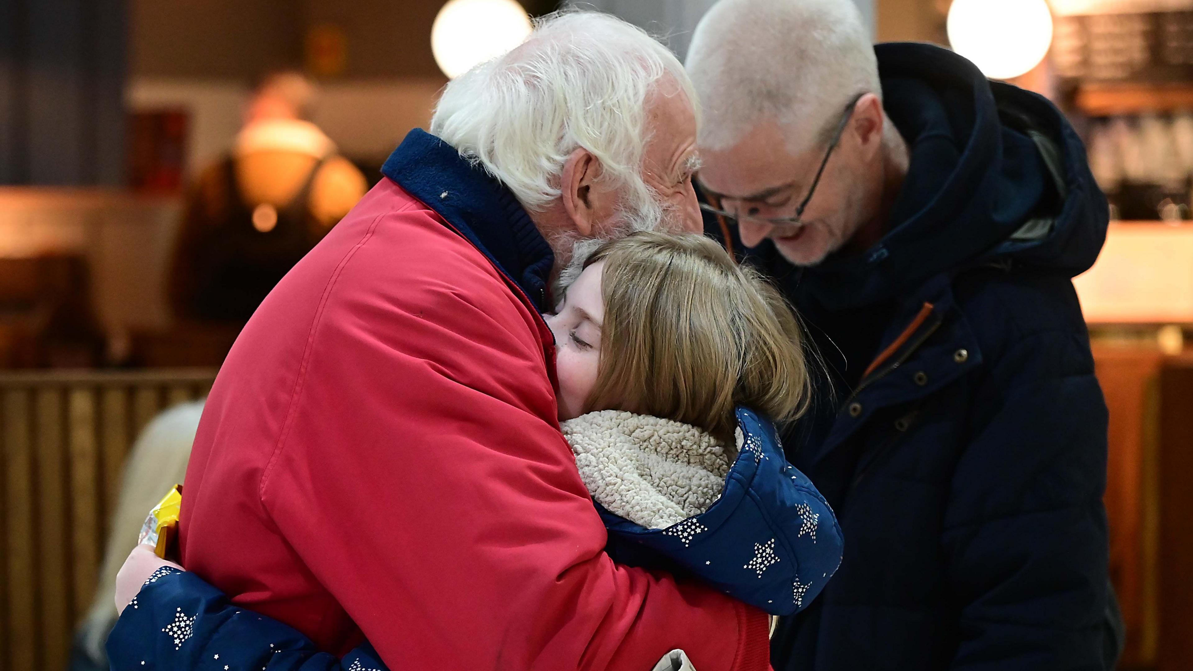 An older gentleman, with white hair and a red coat is hugged by a girl, with sandy hair and a fleece lined coat.