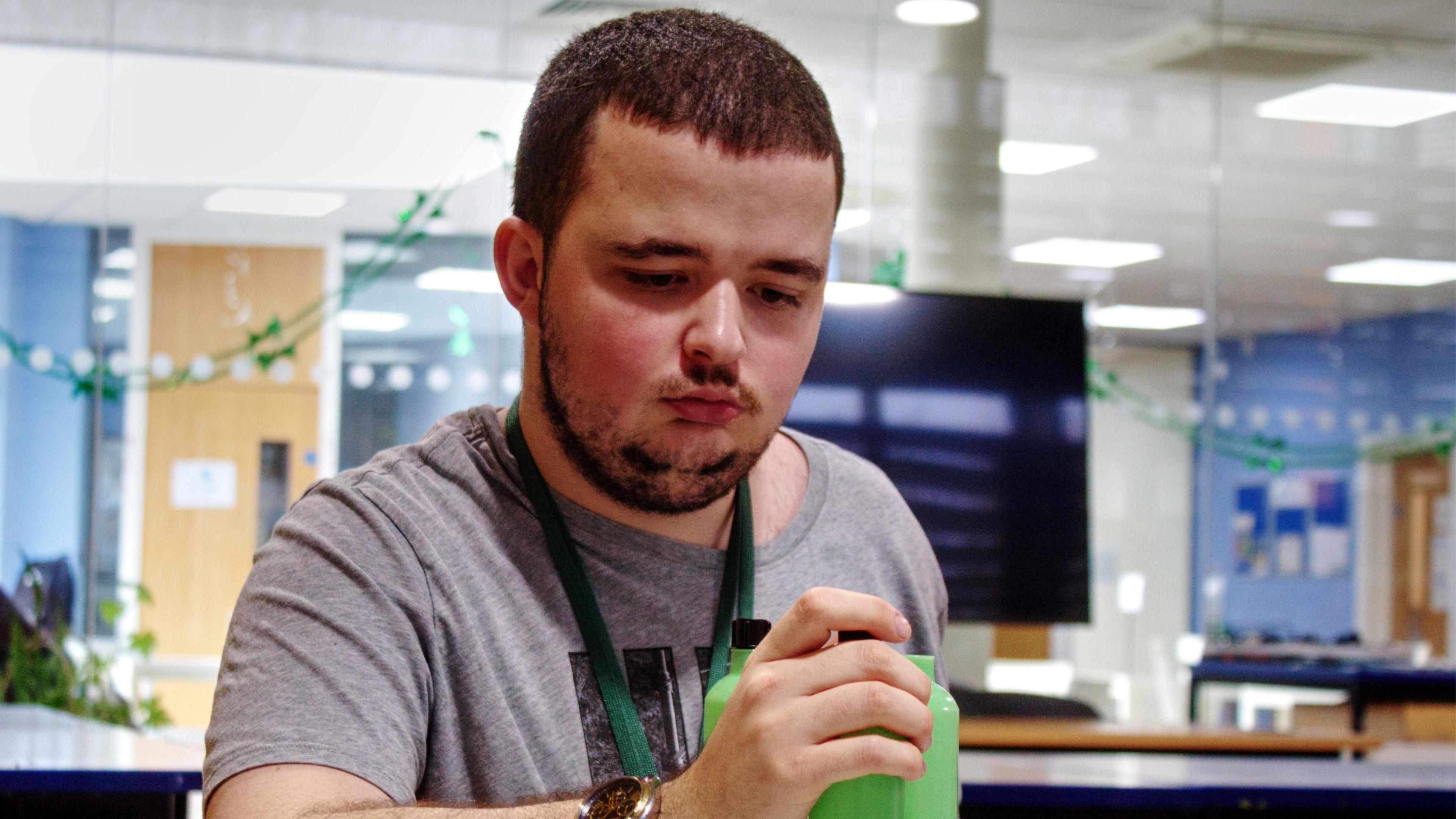 Jack, wearing a grey T-shirt, holding a green time, with a lanyard on. He is in an office, with a monitor on, is looking down, with a slight beard and short dark hair. 