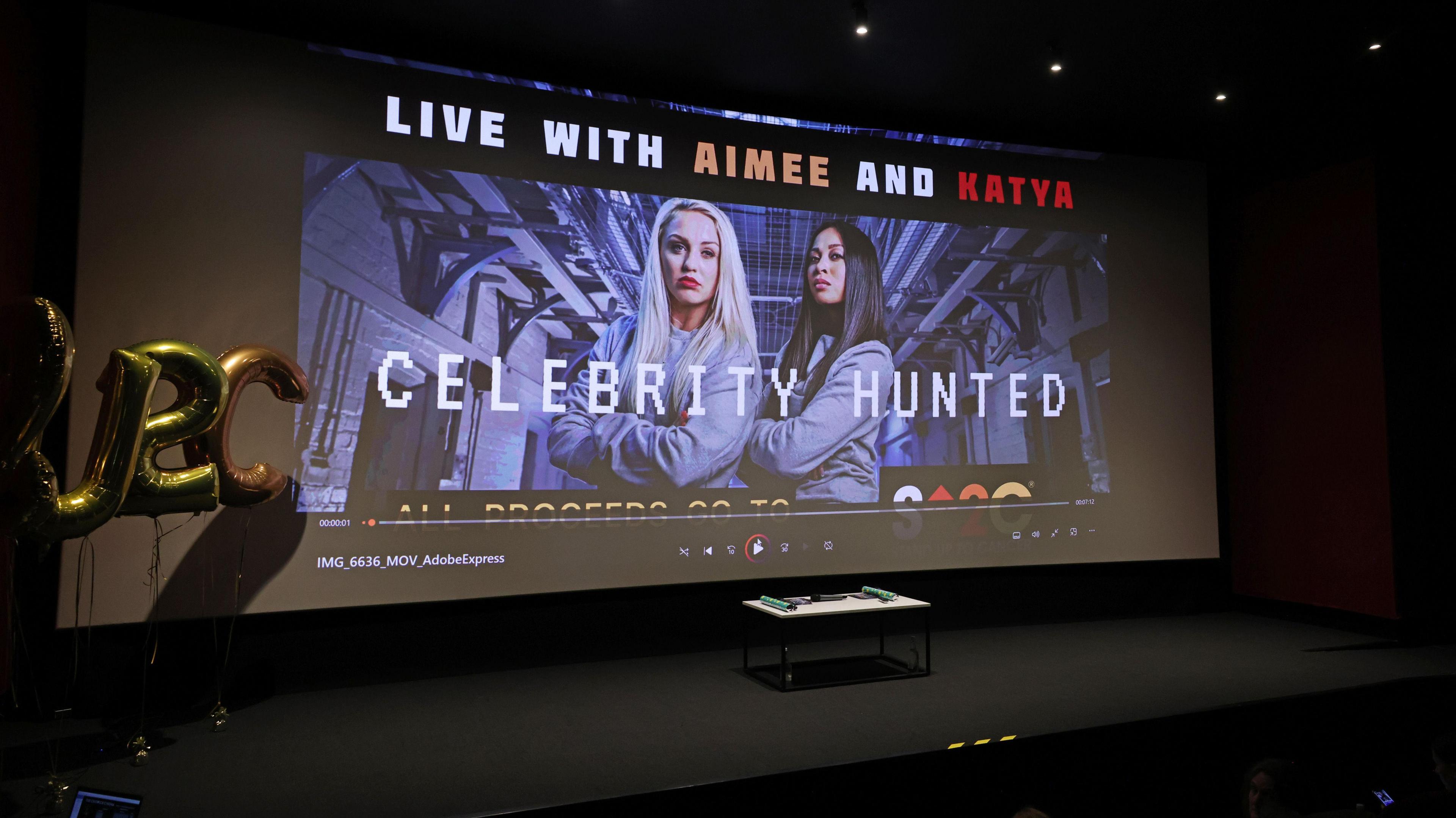 A huge projector says: Live with Aimee and Katya. Celebrity Hunted. There is a stage just below it.