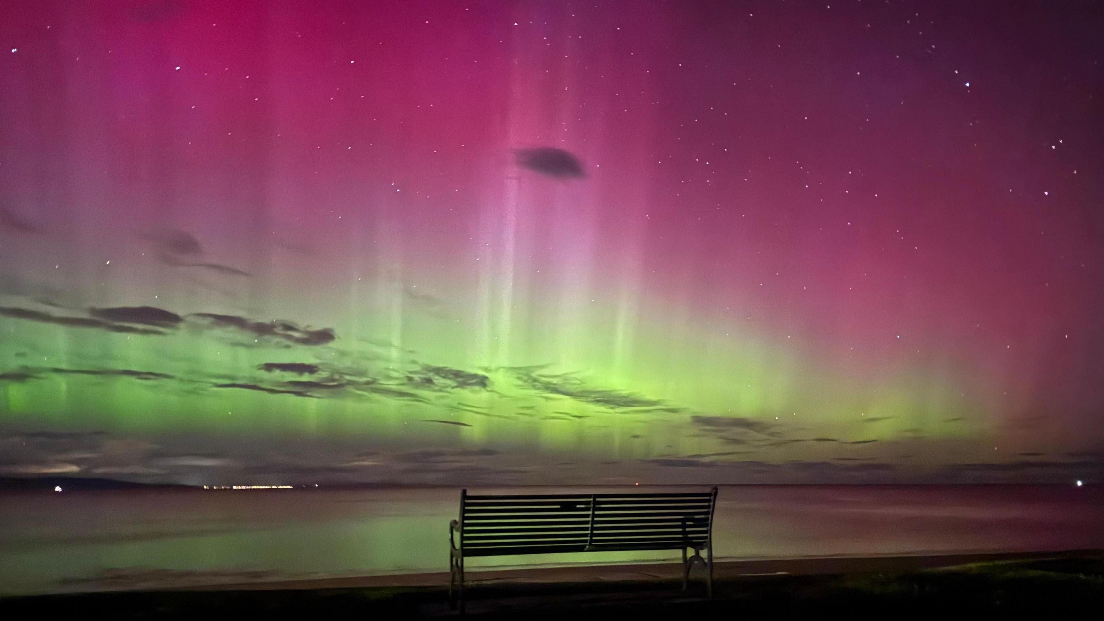 A bench sits in front of the sea in the dark. It is illuminated by a bright green and pink sky, which is dotted with dark clouds. The water reflects from the green lights.
