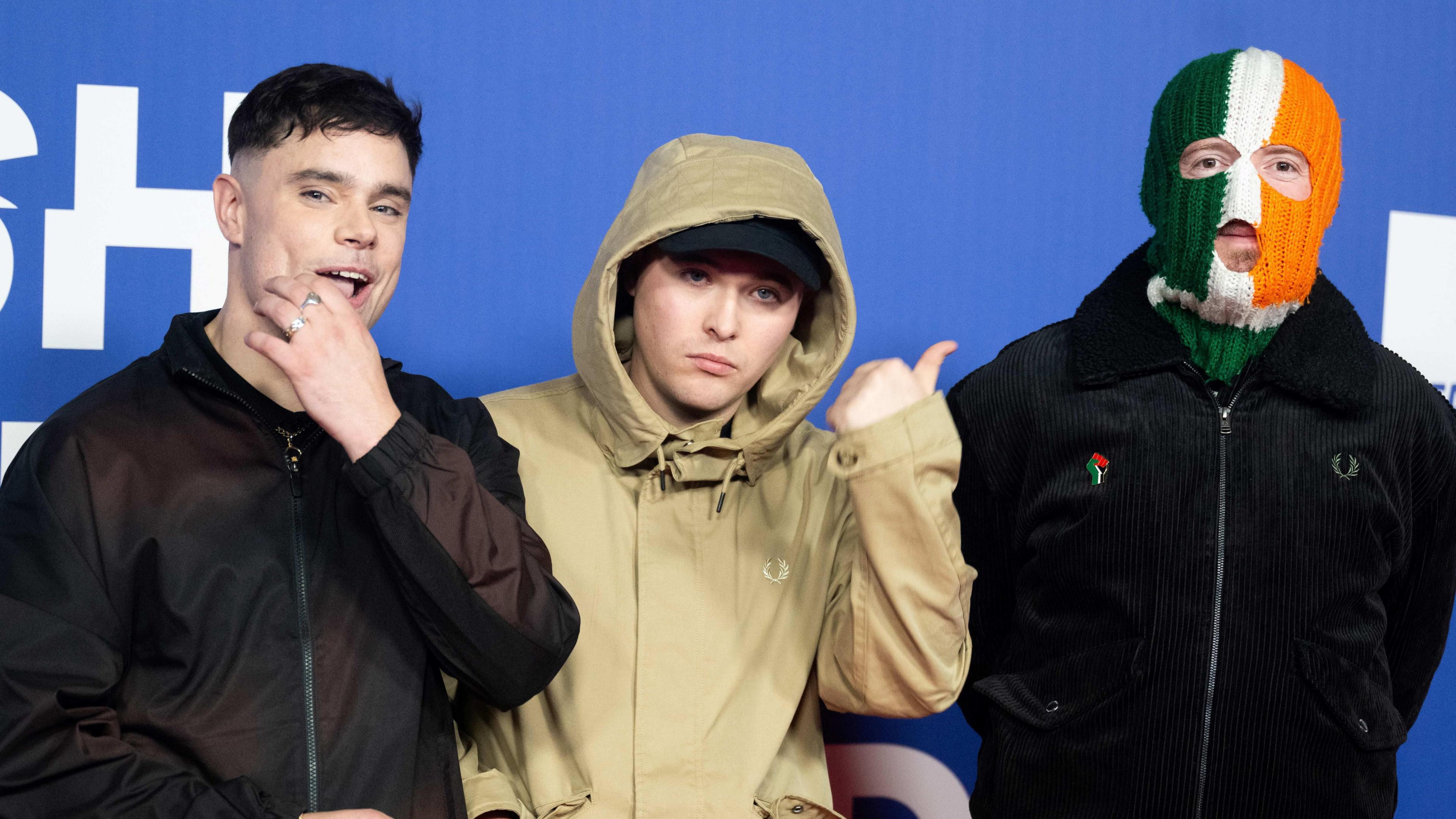 Three young men stand next to each other on a red carpet. One has his hand on his face. The middle man is pointing towards the last man, who is wearing a green , white and orange balaclava over his face. 