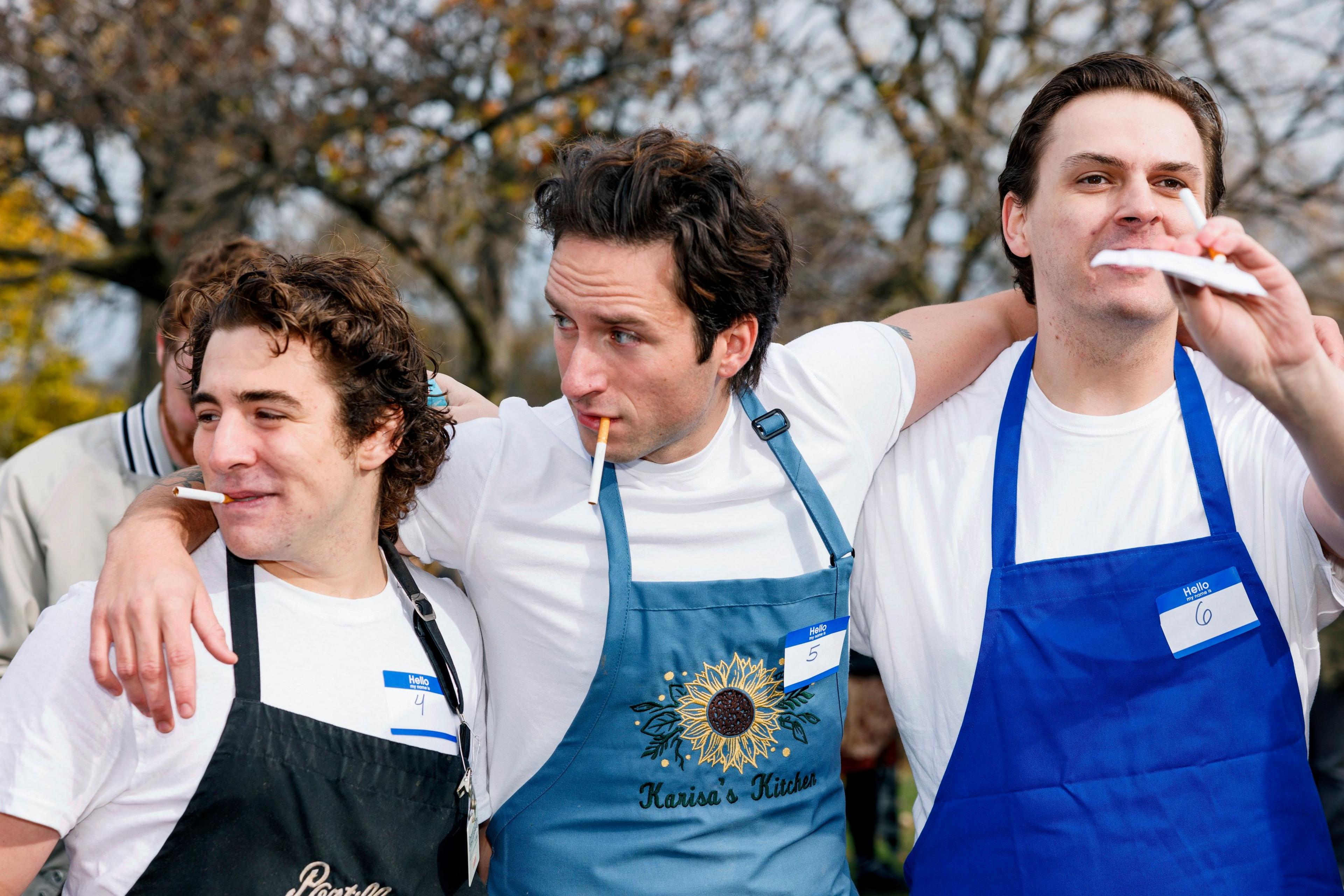 Three Jeremy Allen White lookalikes dressed in chef's aprons and white t-shirts and with cigarettes in their mouths stand, the middle one with his arms over the shoulders of the other two