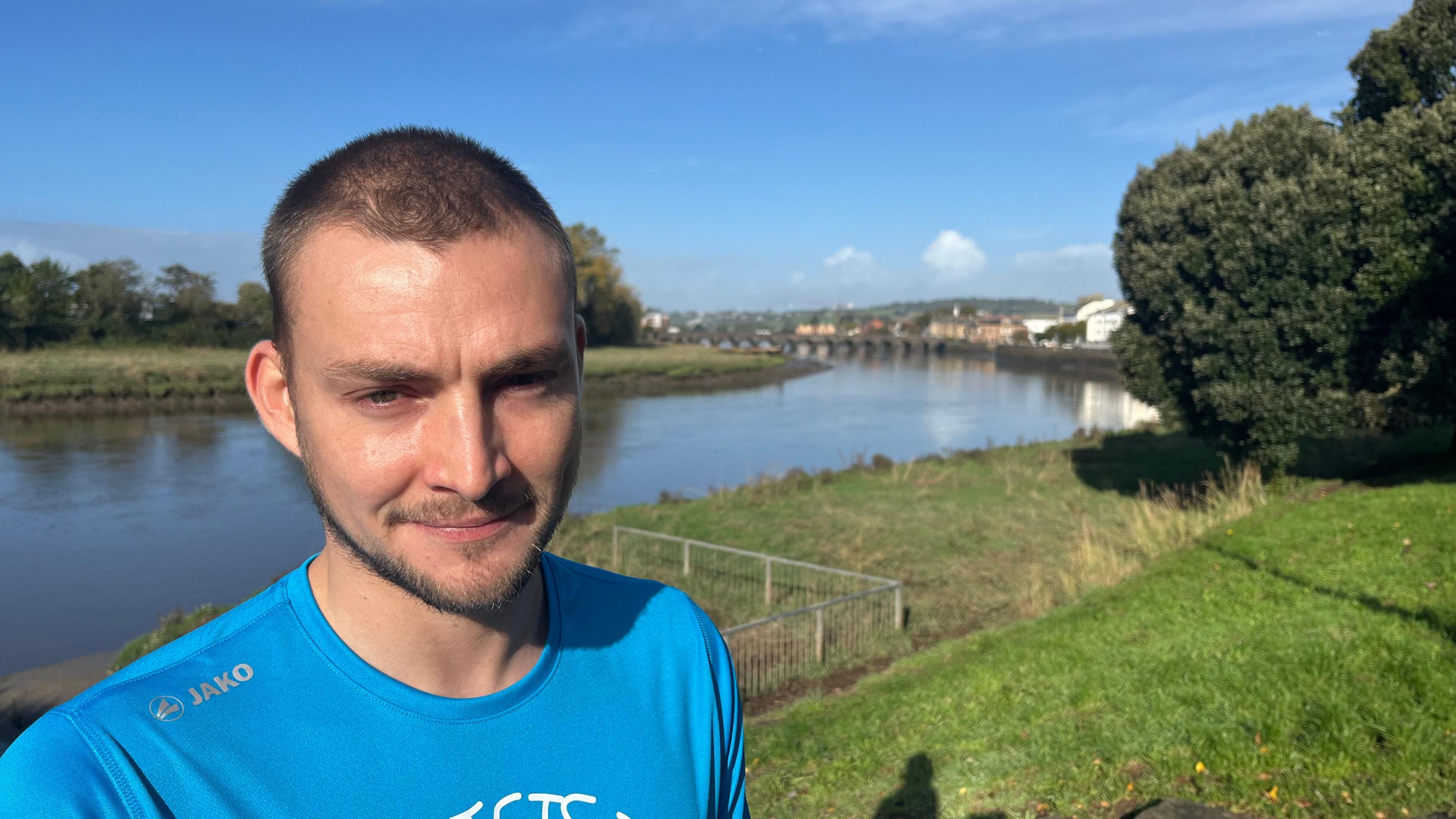 Connor Shannon wearing a blue T-shirt in a park with the river behind him.