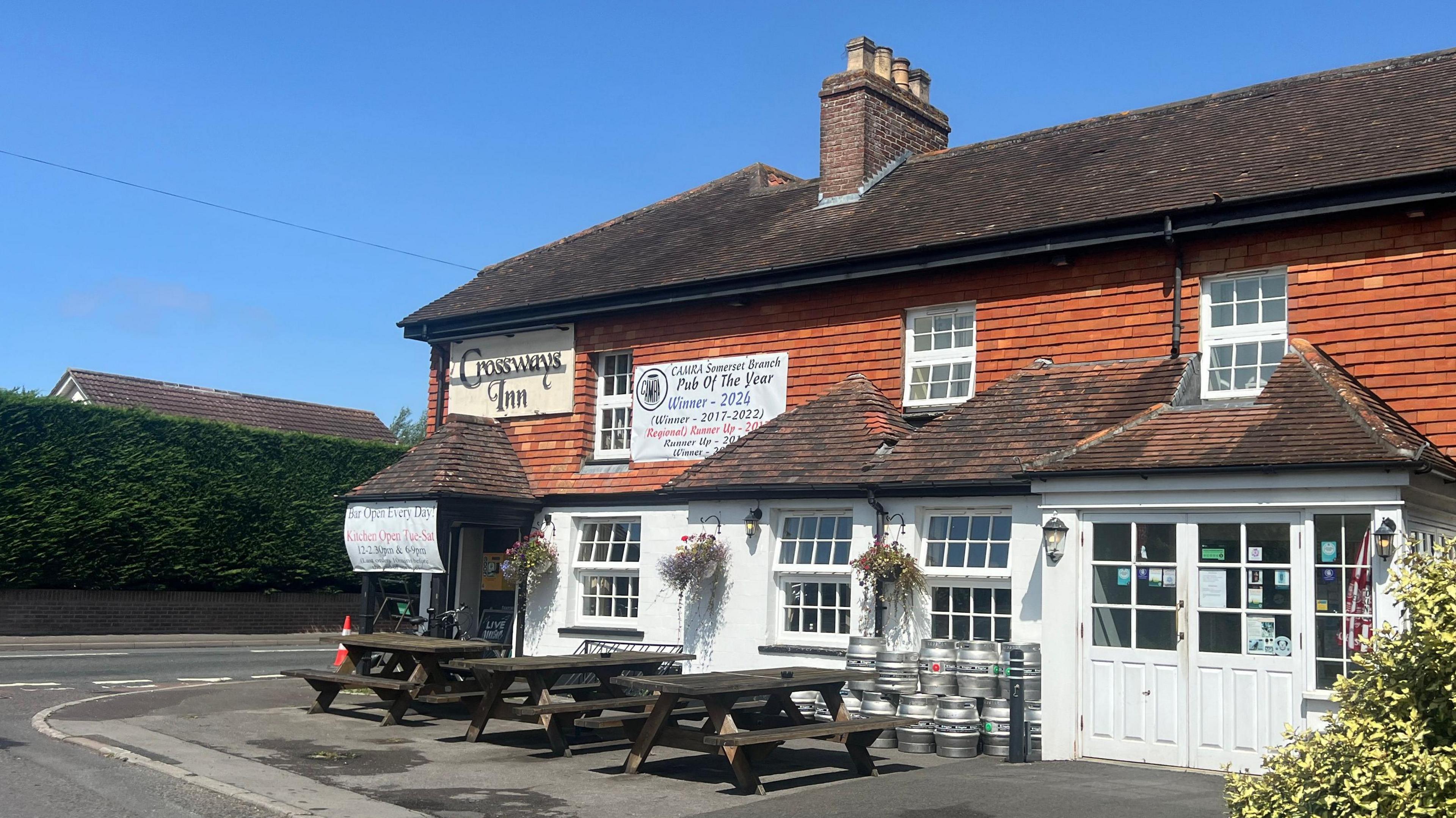 Pub's entrance with picnic benches outside near road junction. The bottom of the pub is white with orange bricks in the middle and a black roof.