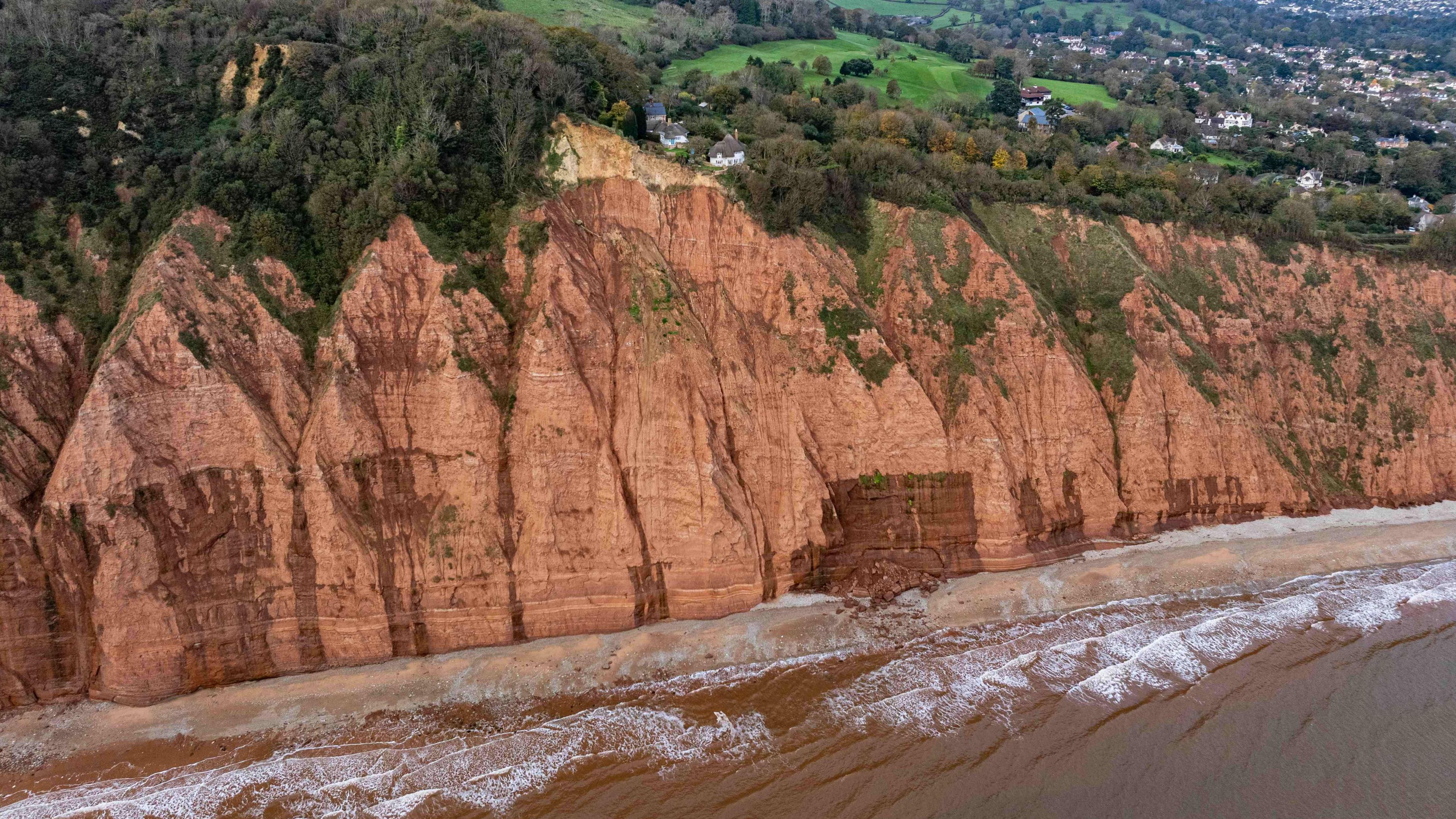 A house with a conical roof sits close to the edge above the sea with more houses further inland and nearby hedges and trees along with signs of a cliff fall nearby with mud going into the sea.