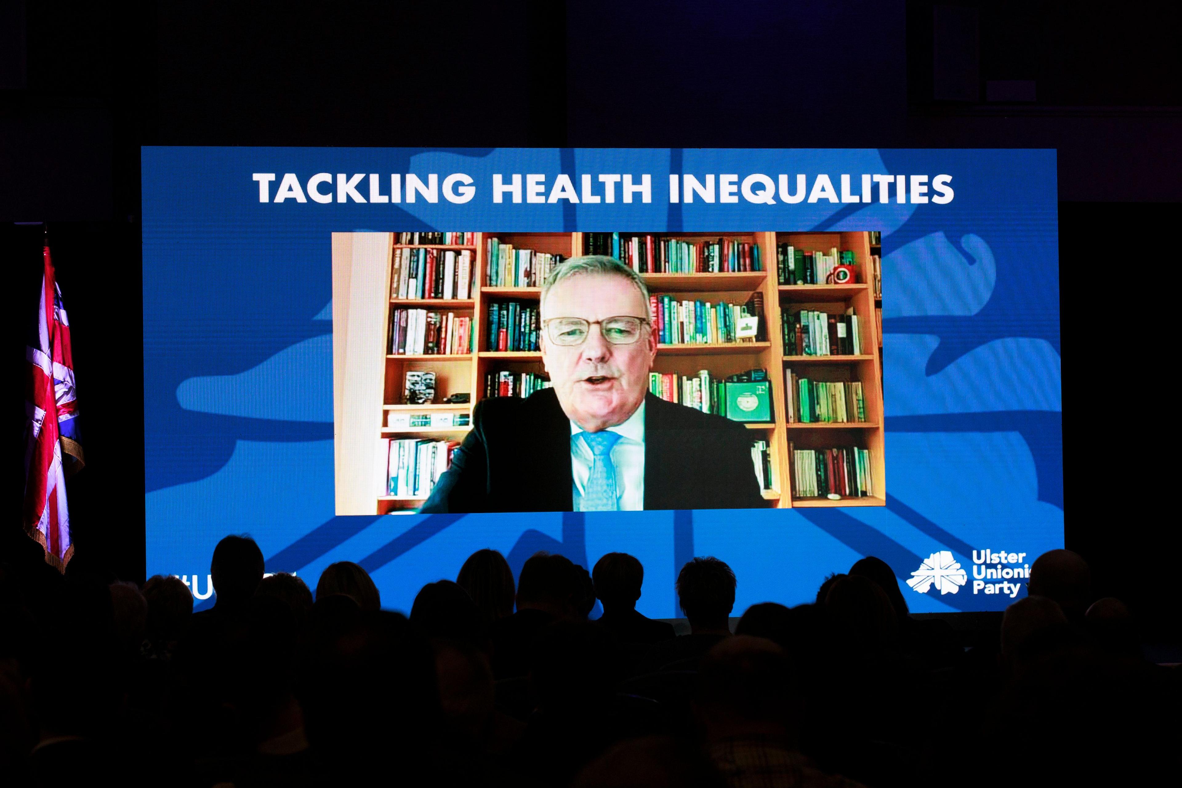 Ulster Unionist leader Mike Nesbitt on a screen at the party conference making his address remotely. Above the video it says 'Tackling Health Inequalities'. There is a Union Jack hanging beside the screen.