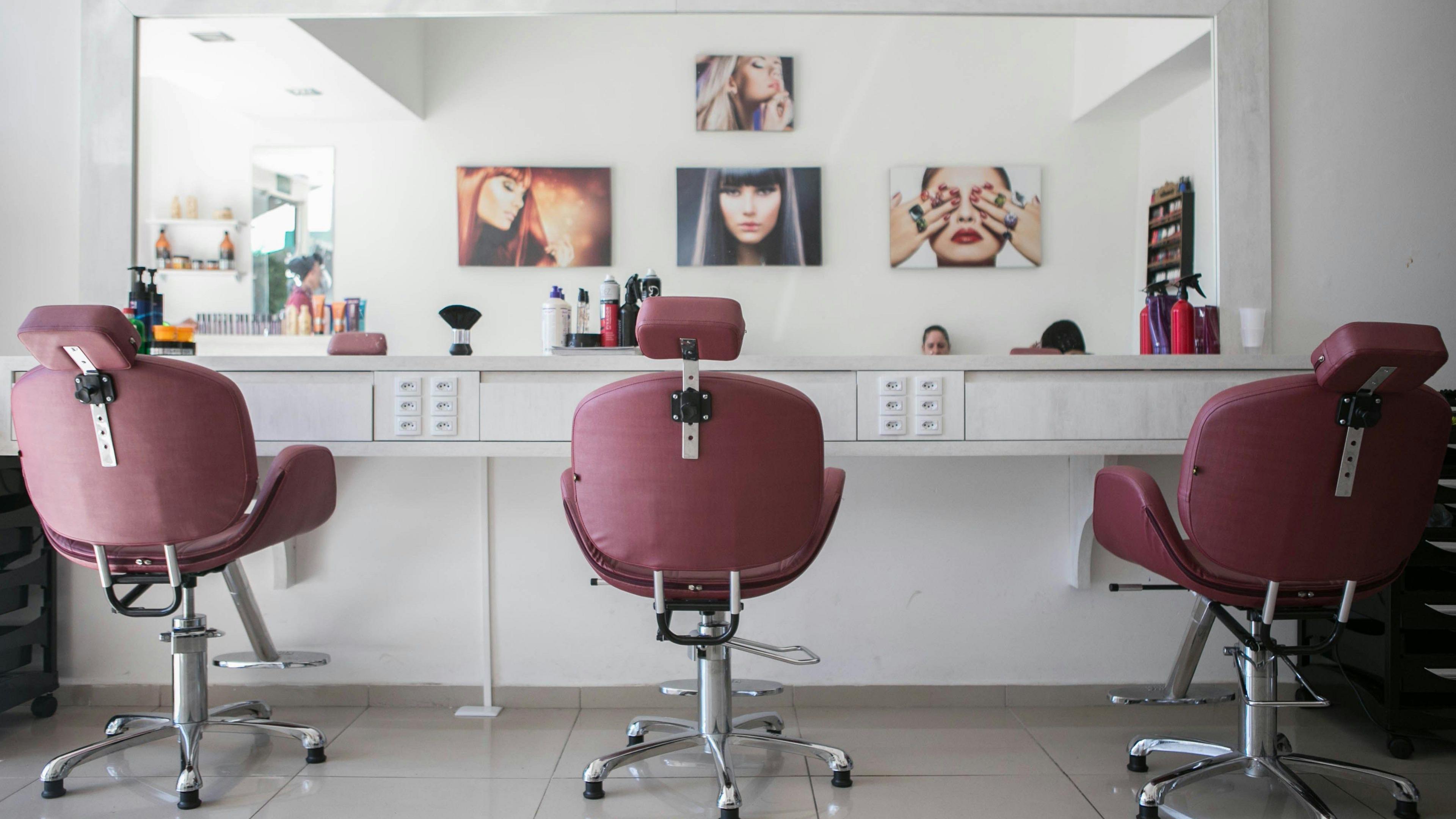 A stock image of an interior of a white beauty salon with a tiled floor. Three wine coloured swivel chairs are facing a mirror. Four paintings of women's faces can be seen in the reflection. Various hairspray bottles and makeup equipment are on the table below the mirror.