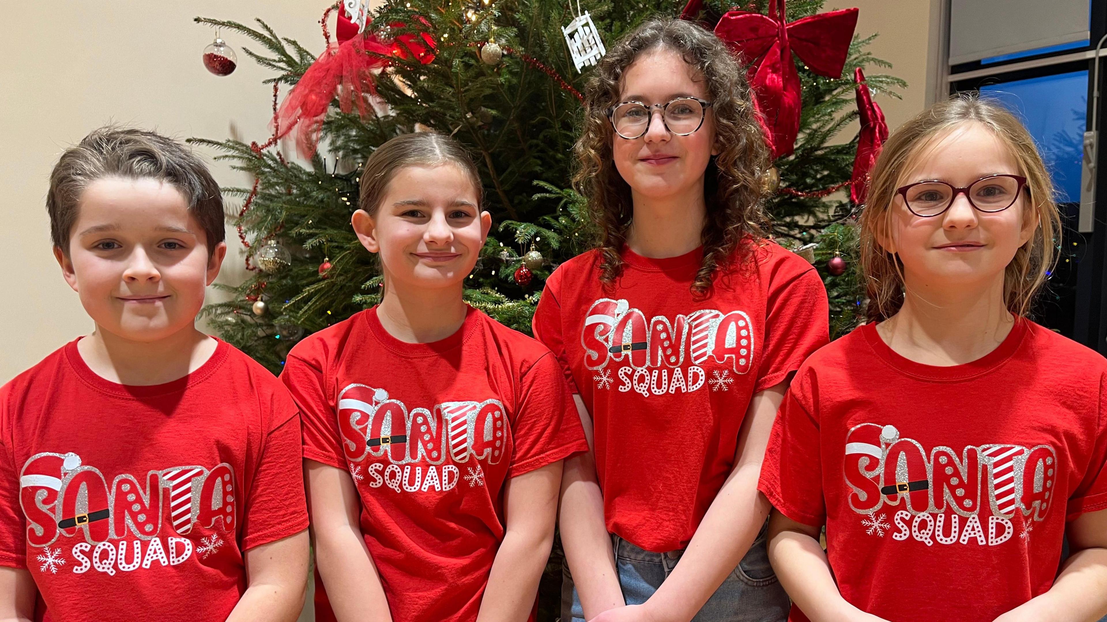 Four young children stand in front of a Christmas tree. They are wearing red T-shirts with Santa Squad written on them.