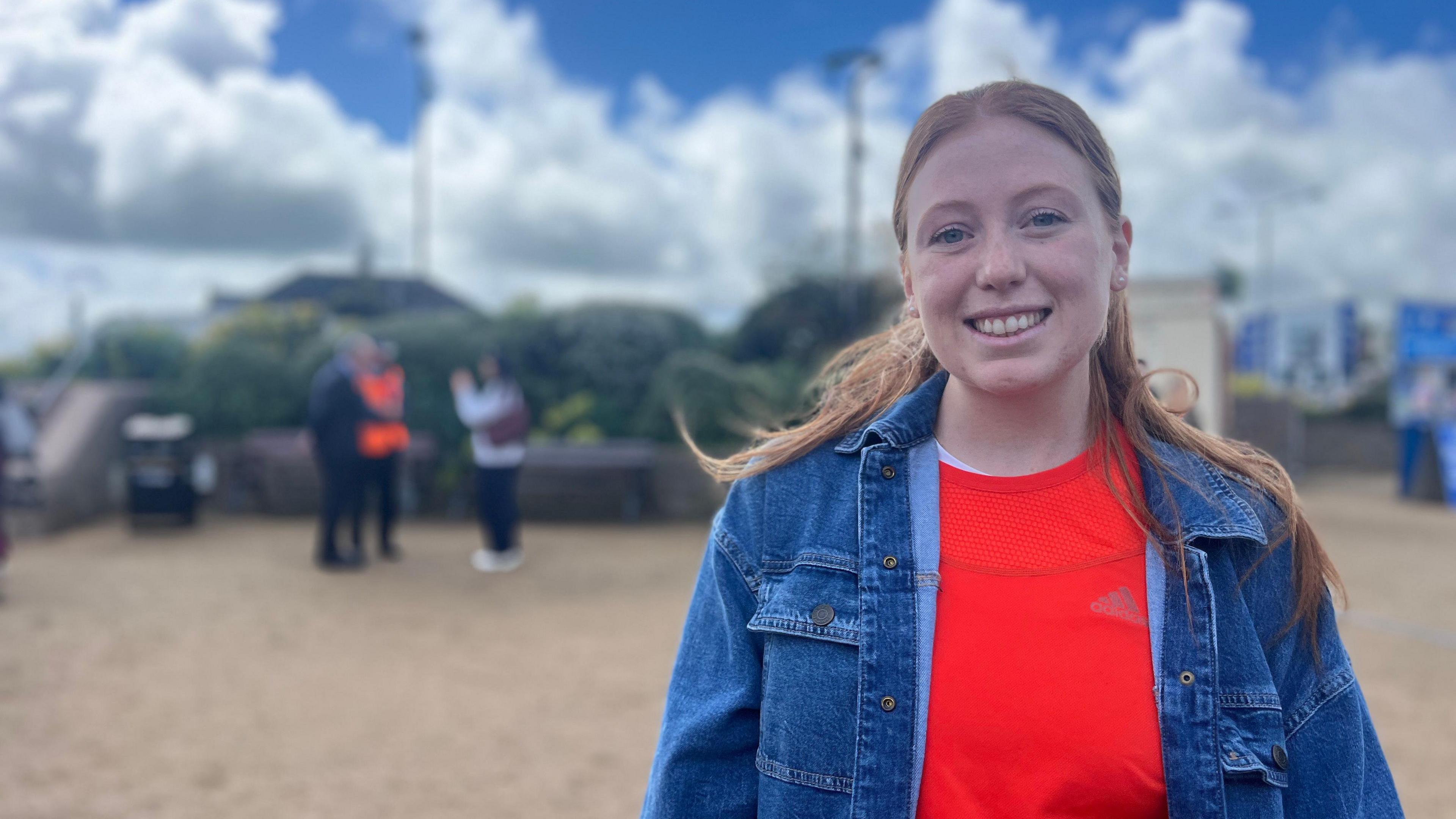 Adele smiles at the camera as a few people talk behind her. She has a denim jacket on with a orange sports top underneath. Adele has long ginger hair which comes down to her shoulders.