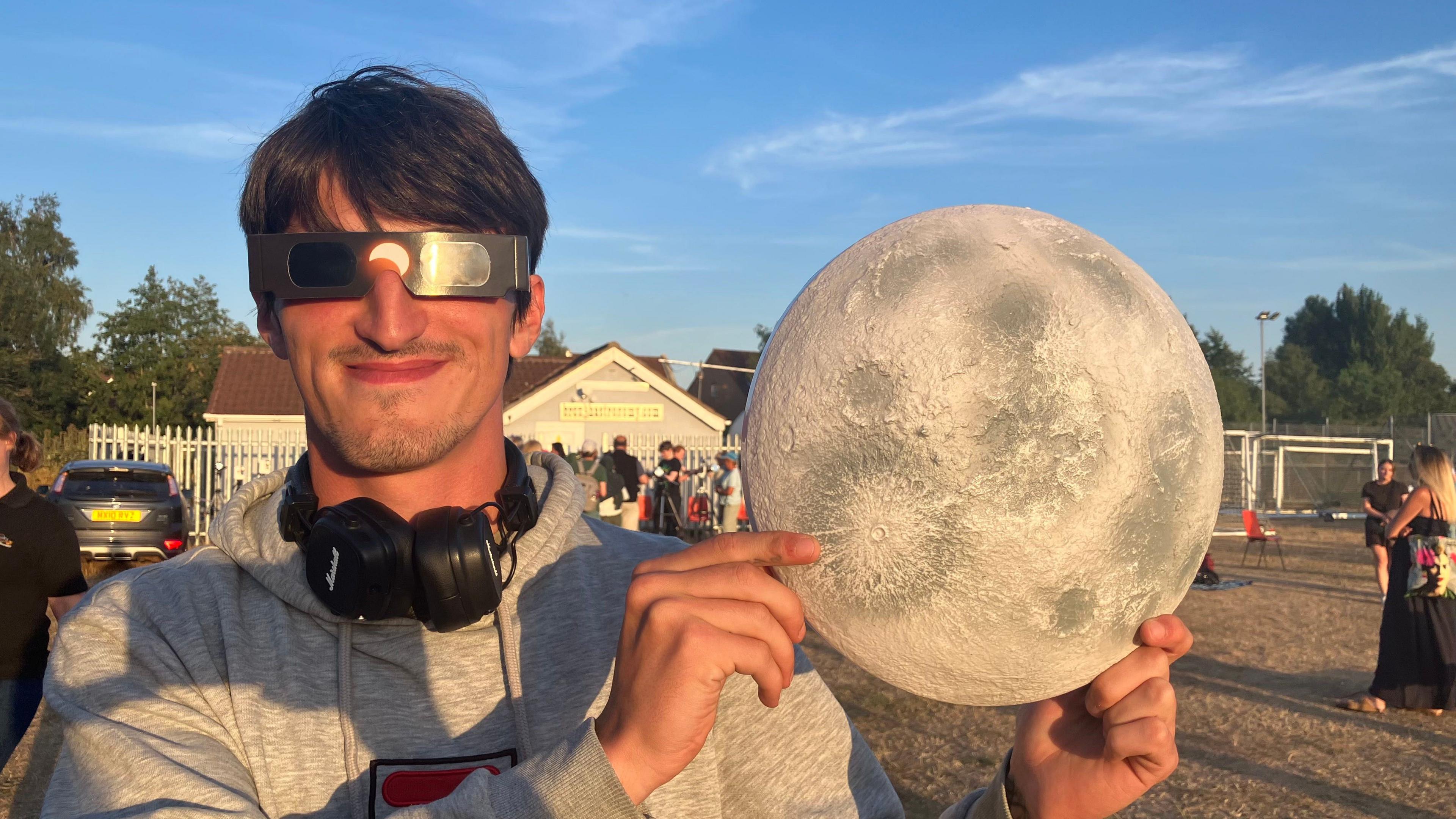 Kian Lodge smiling at the camera, holding a toy Moon. He is wearing black eclipse glasses, black headphones around his neck and a grey hoodie. He is standing in a field. The sky is blue.