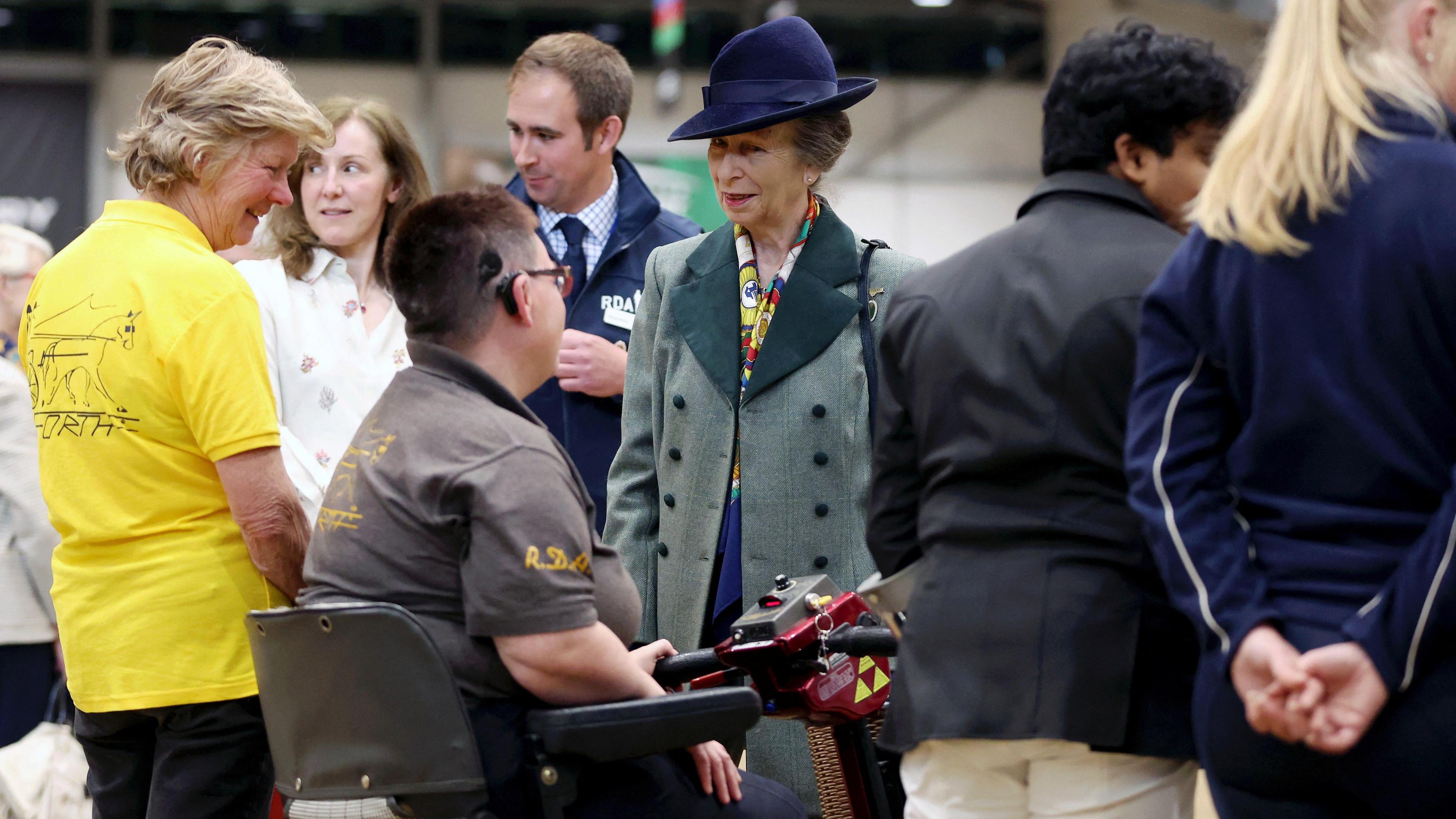 Princess Anne chatting with people at an event. She is wearing a hat and green coat and smiling. 